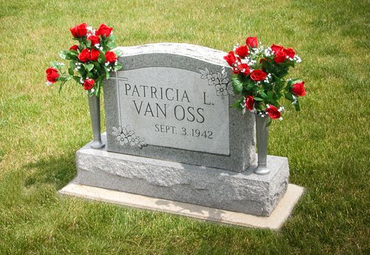 Gray gravestone with red flowers on a green lawn, engraved “Patrick L. Van Oss.”