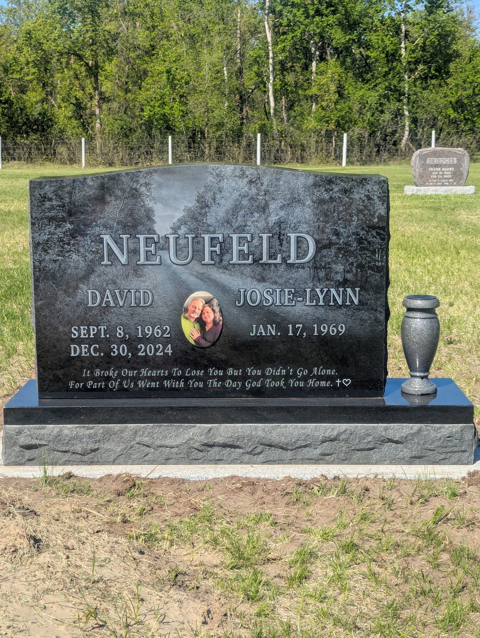 Black granite memorial headstone in a cemetery, with etched name and dates and a small vase beside it.