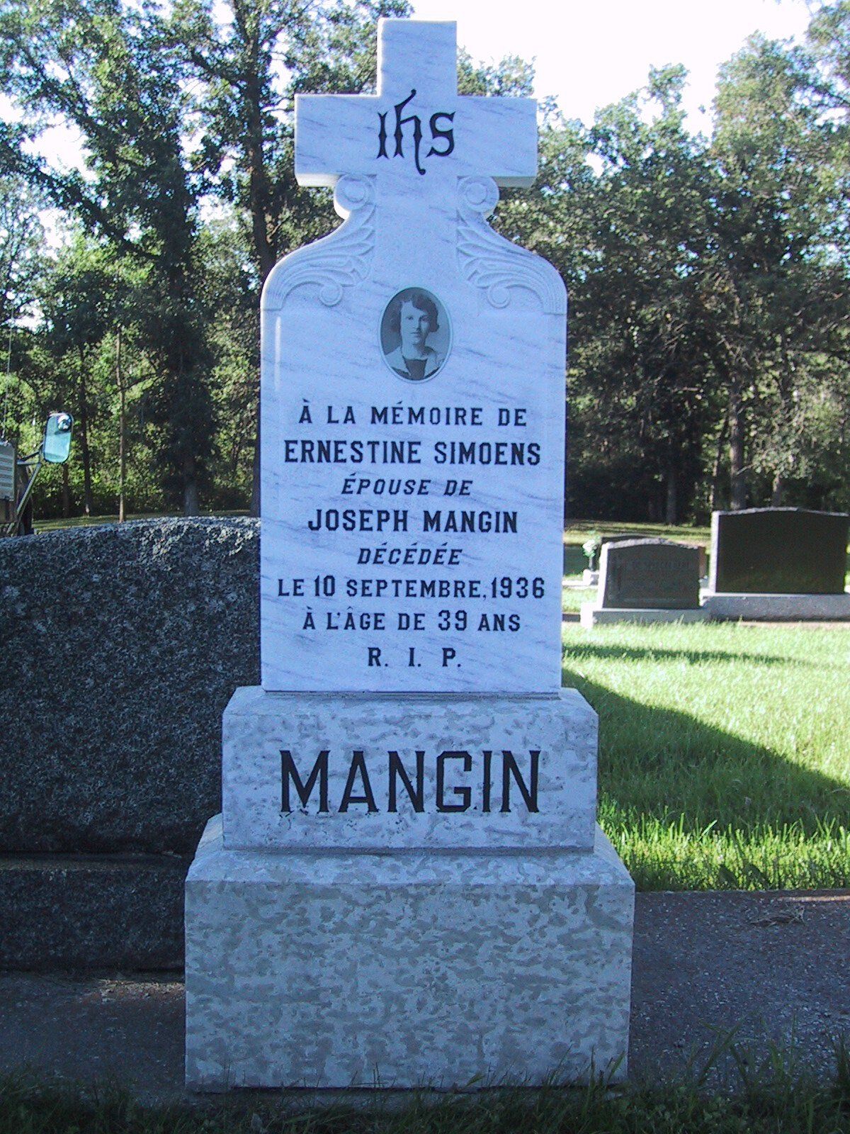Gray stone memorial cross in a cemetery, engraved “MANGIN,” surrounded by trees.