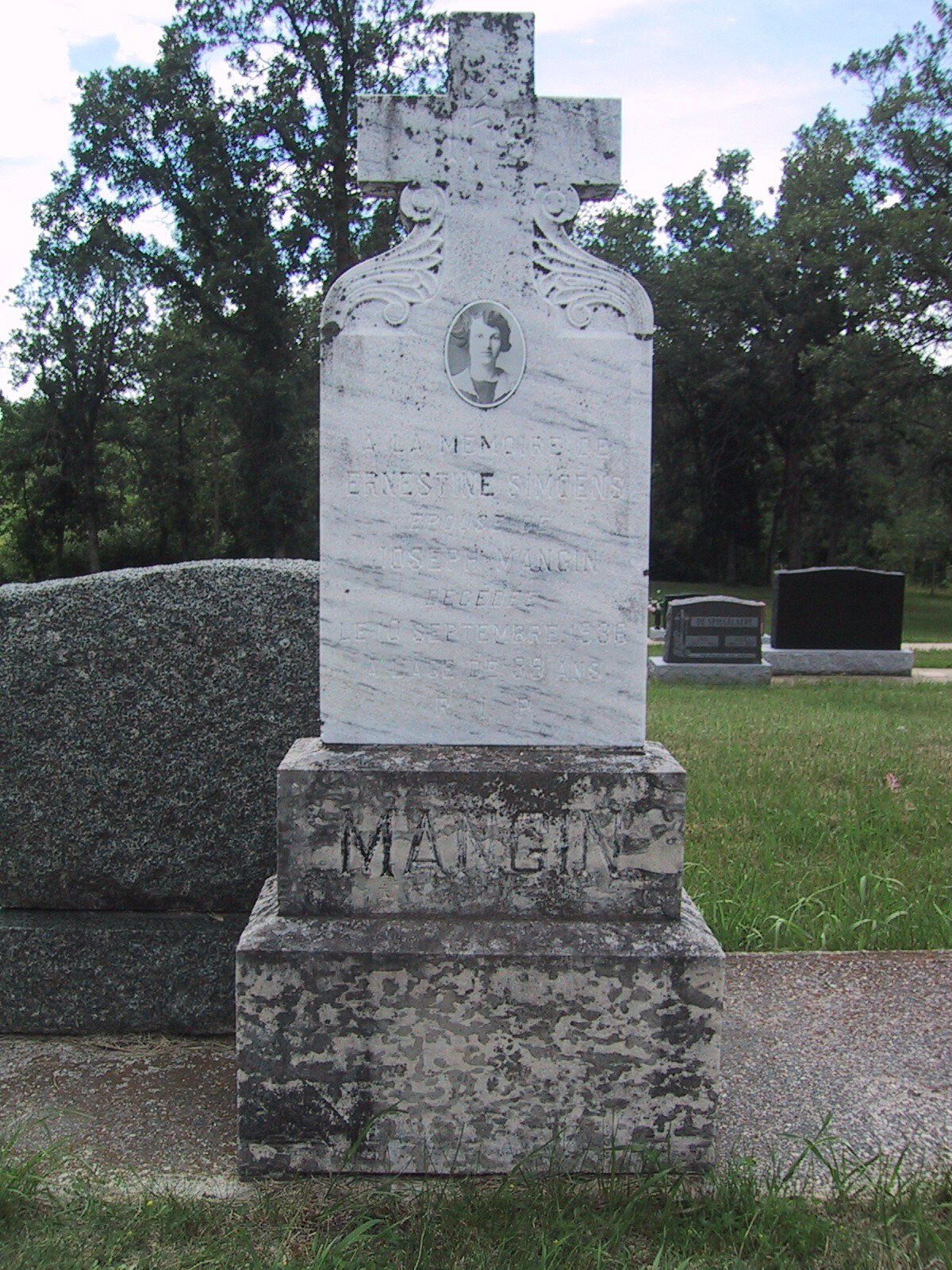 Weathered white gravestone with a cross in a grassy cemetery, framed by trees.