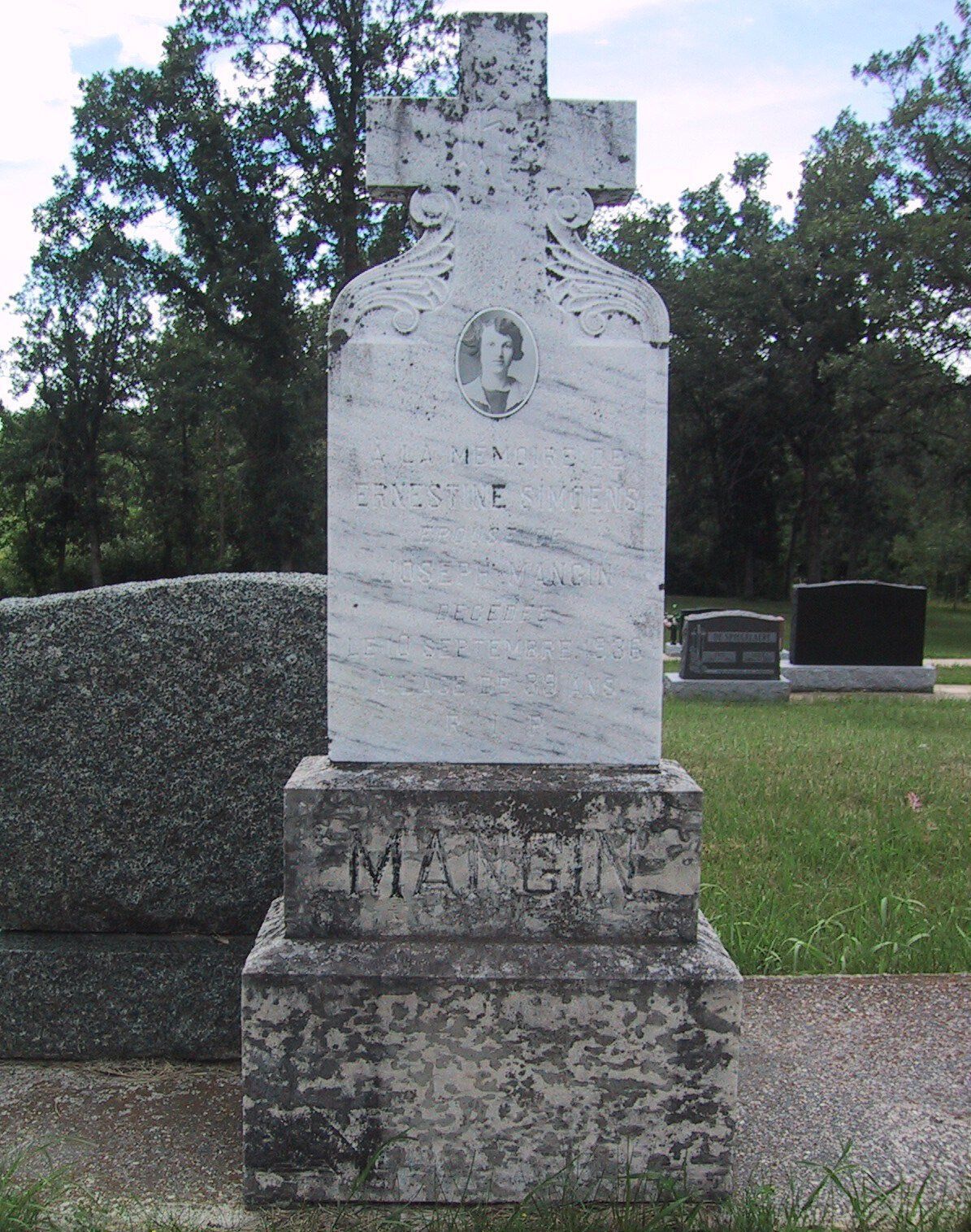 Weathered white gravestone with cross-shaped top in a grassy cemetery, trees in background