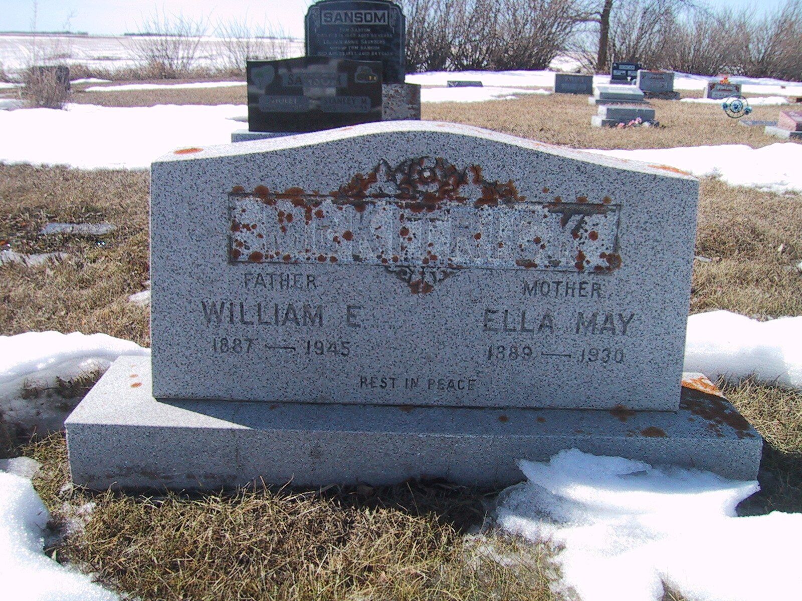 Snow-covered cemetery gravestone with weathered inscription and a smaller marker behind it