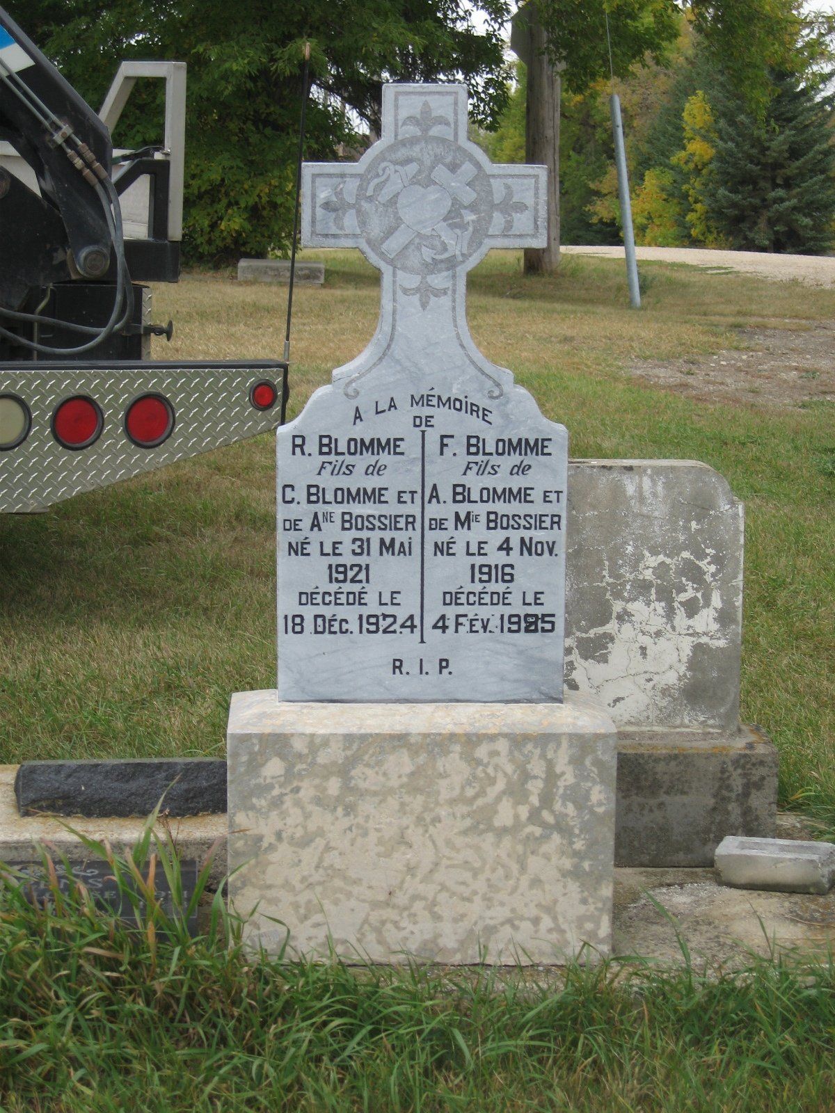 Weathered gravestone with carved cross in a grassy cemetery, beside a vehicle tire on the left.