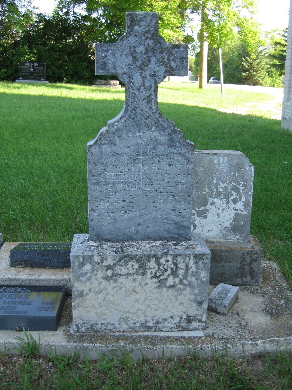 Weathered gravestone in a grassy cemetery, with a cross-shaped top and faded inscription.