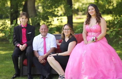 Four people posing outdoors; one wears a bright pink ball gown.