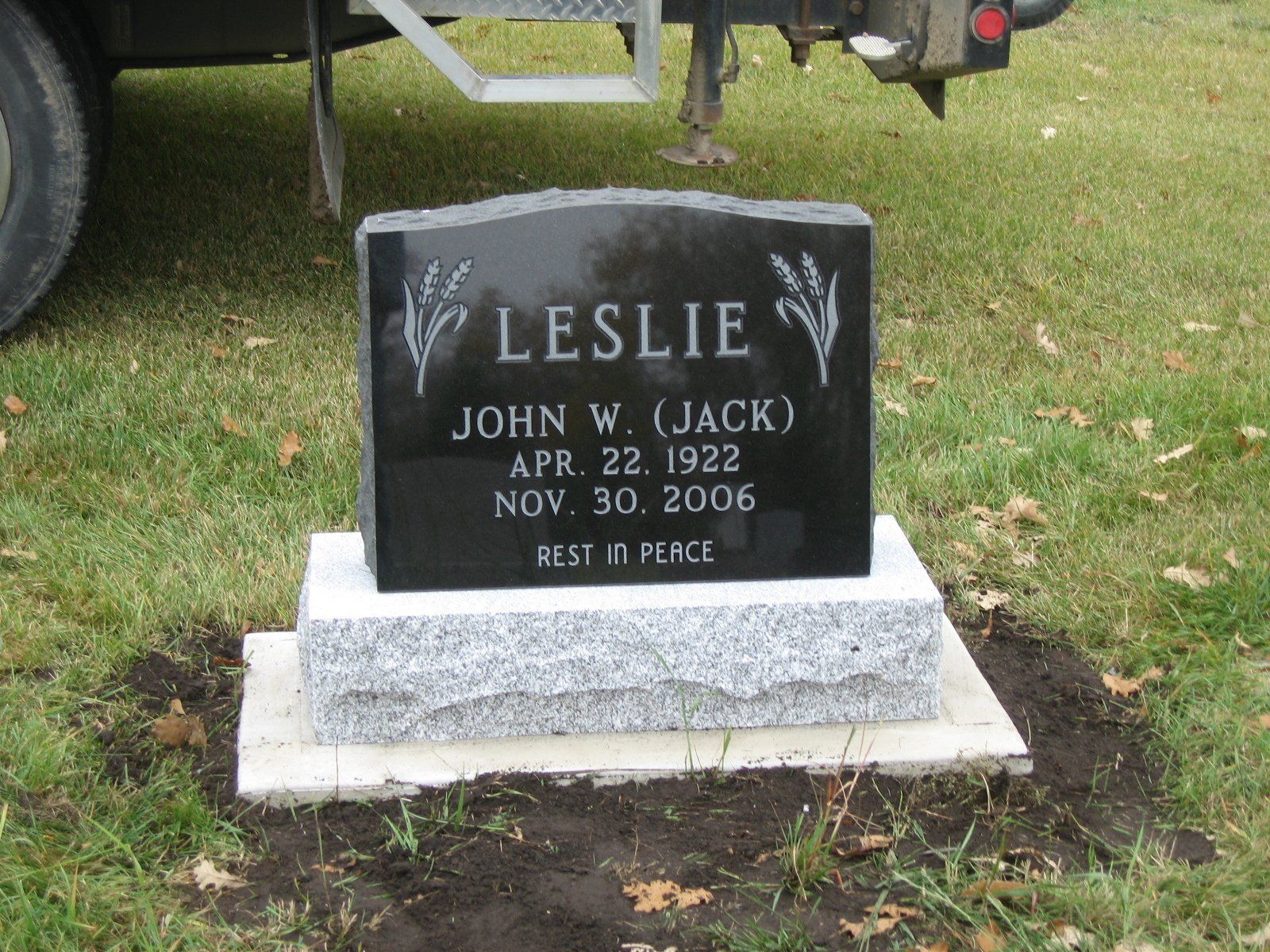 Black gravestone for Leslie John W. Jack with dates, set on a grassy lawn beside a vehicle