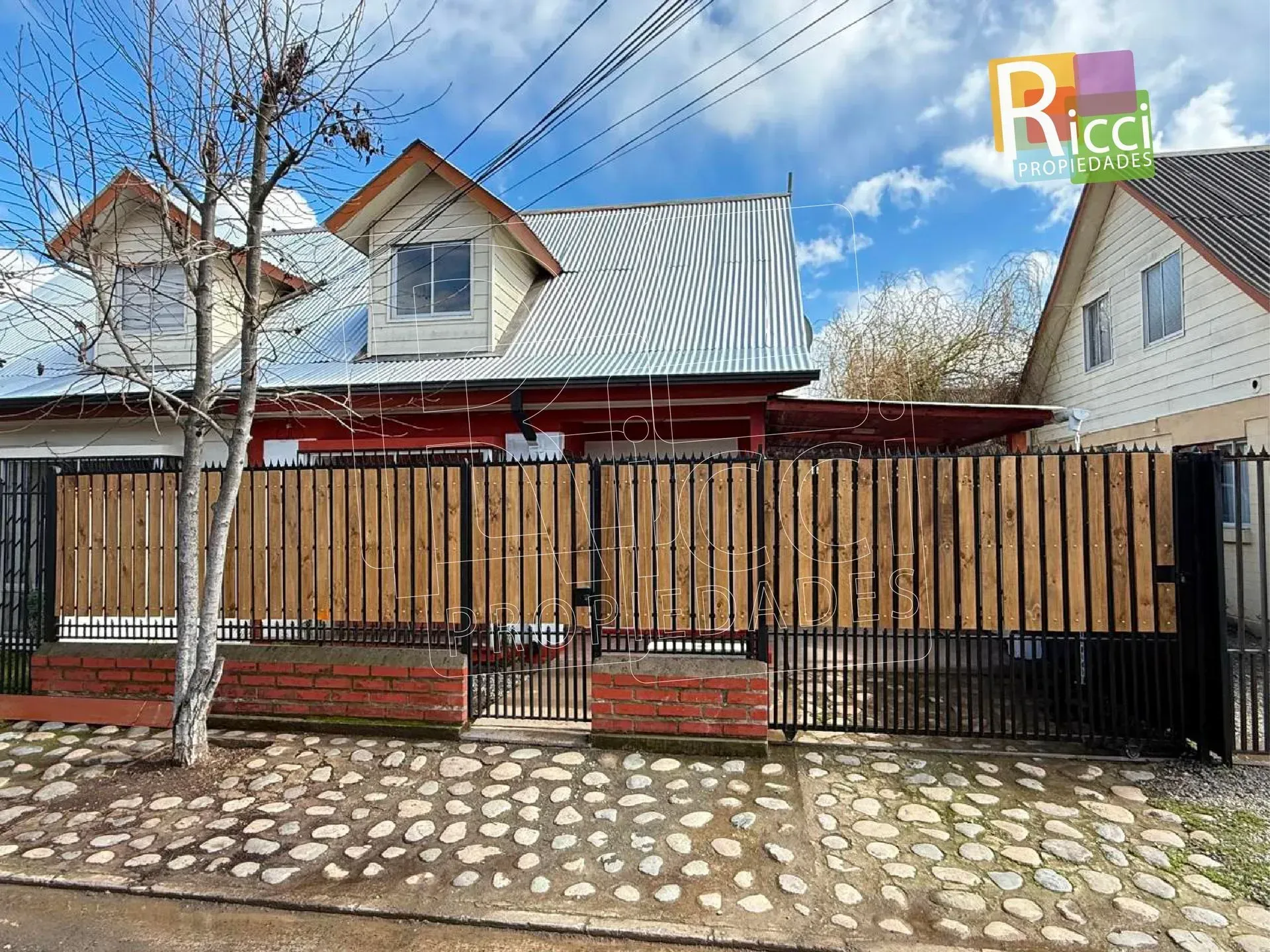 Casa de madera con techo de metal, jardín delantero cercado y entrada de adoquines bajo un cielo azul