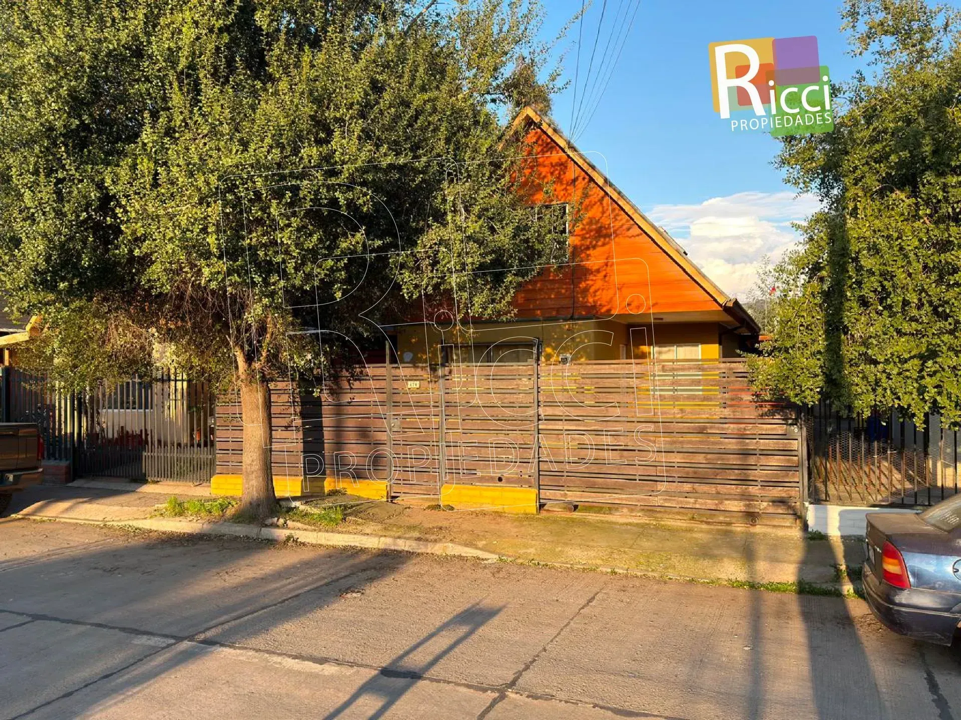 Casa de madera con techo naranja detrás de un árbol, junto a una calle en un día soleado