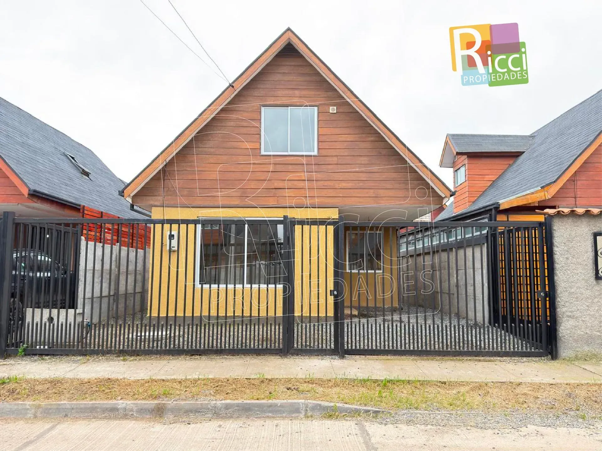 Vista frontal de una casa pequeña con revestimiento de madera, paredes inferiores amarillas y una cerca y un portón de metal negro