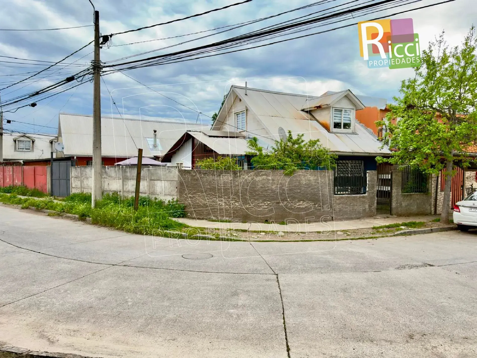 Esquina de una calle con un muro bajo de hormigón, casas, postes de servicios públicos y árboles bajo un cielo nublado