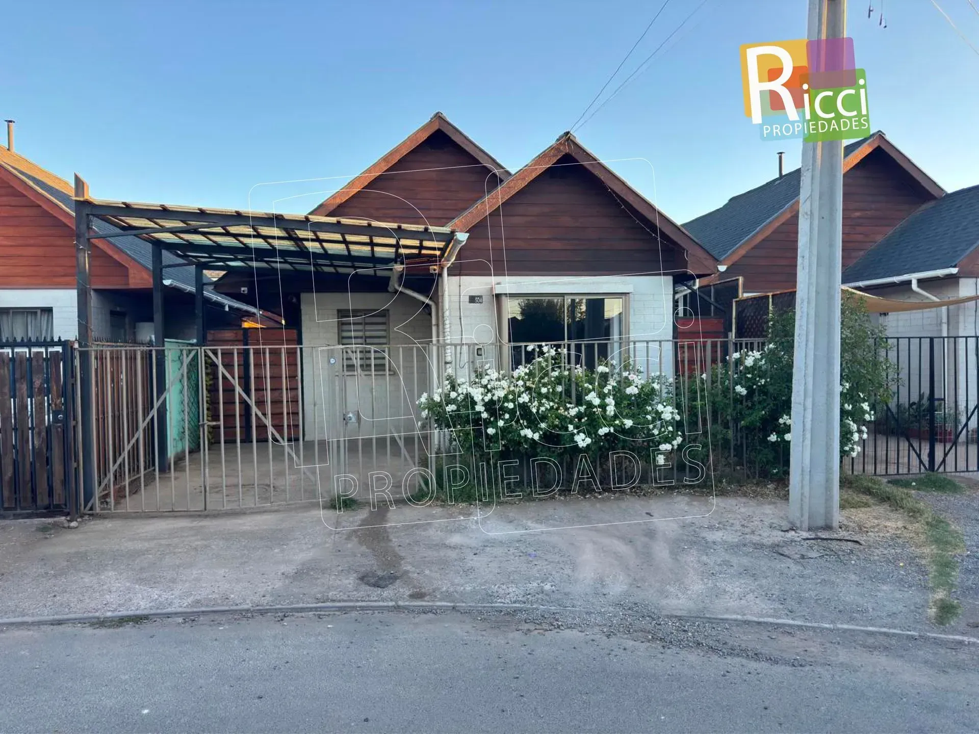 Casa de una sola planta con portón metálico, cochera y arbustos de flores blancas a lo largo de la cerca del frente.