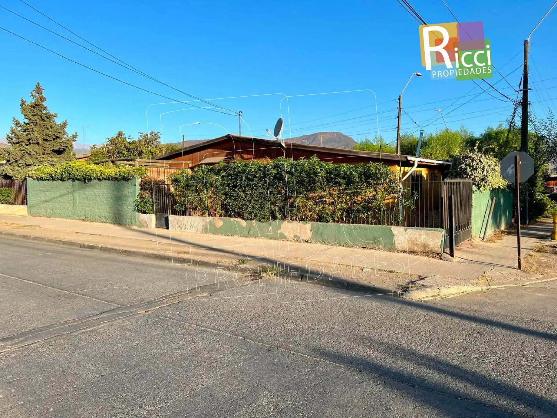 Esquina de una calle con una casa baja detrás de una valla verde, bajo un cielo azul despejado