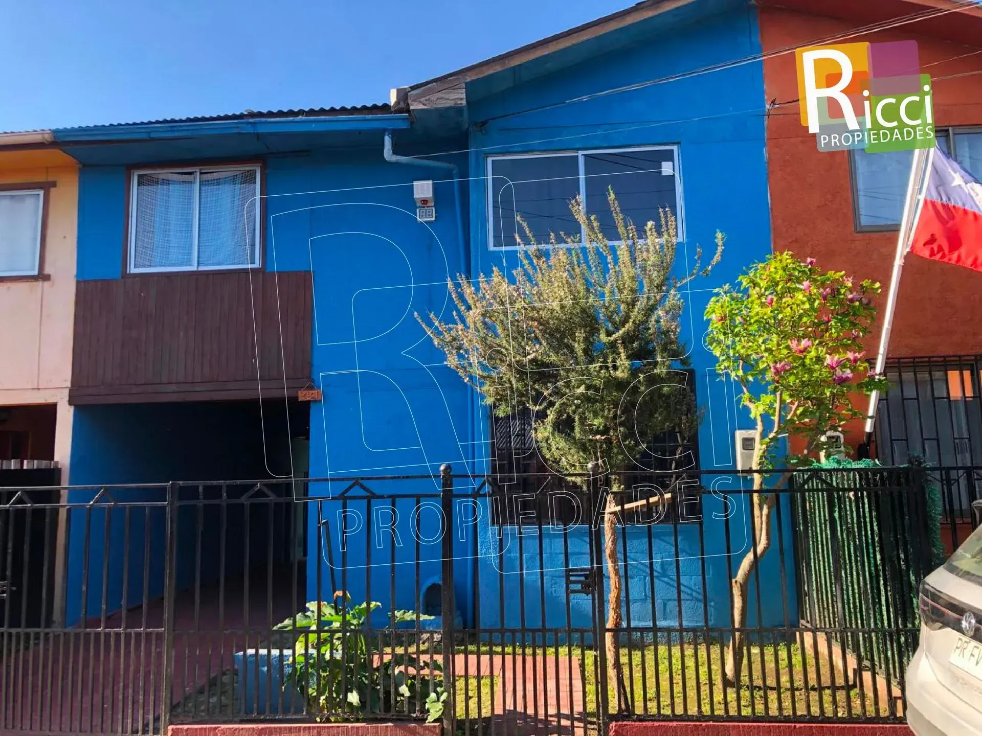 Casa azul de dos plantas con una cerca negra, árboles delante y un letrero verde junto a la entrada