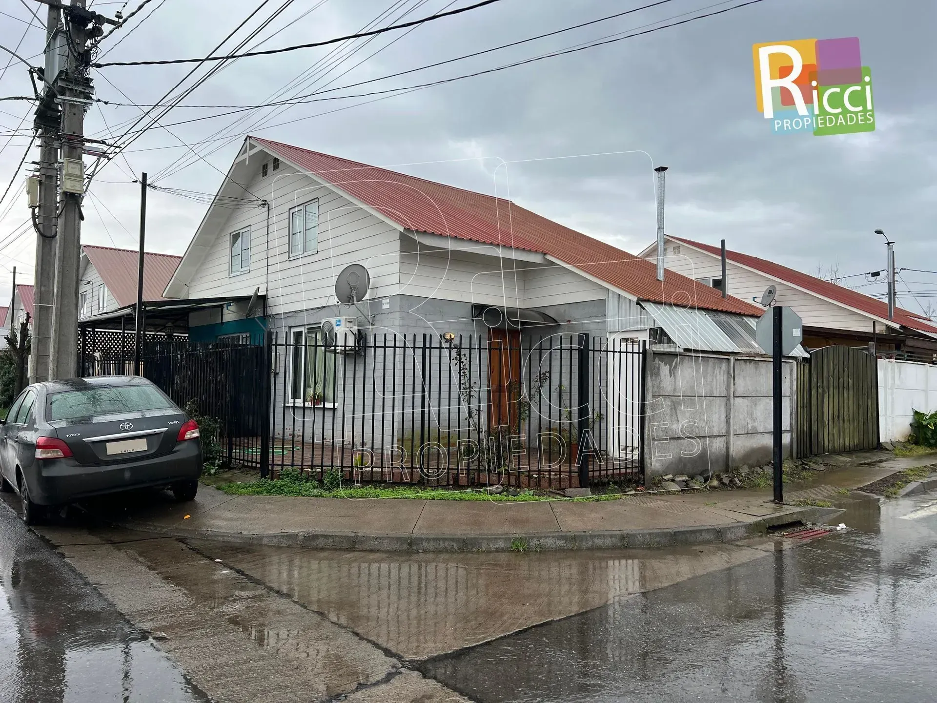 Esquina lluviosa con una casa blanca, tejado rojo, coche negro y una valla junto a la acera mojada.