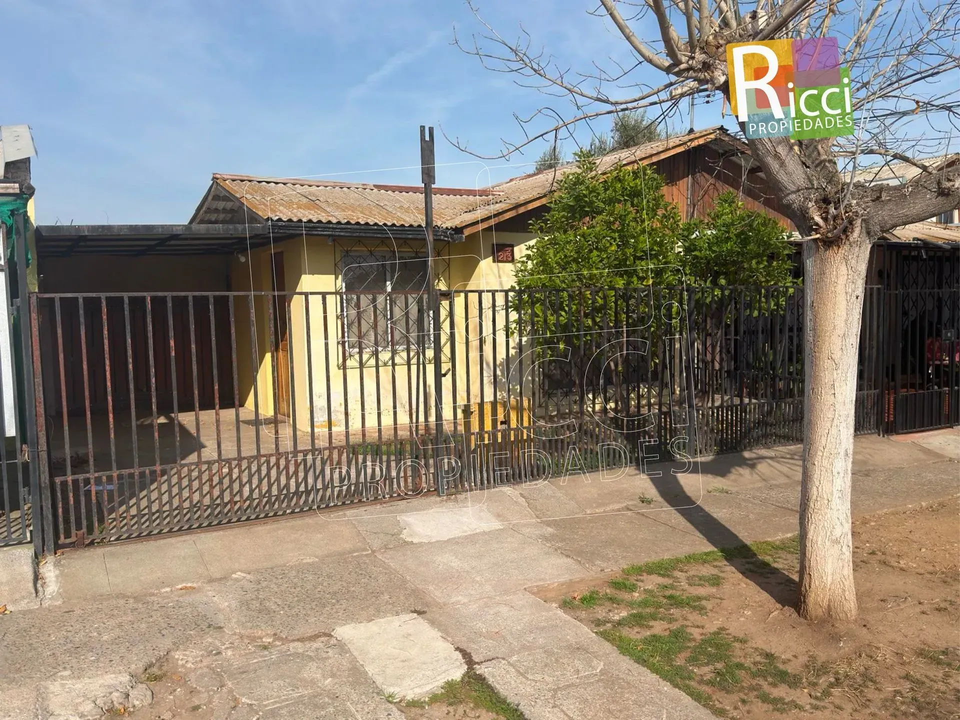 Casa de una planta con una valla metálica, un árbol y una acera pavimentada en una calle soleada.