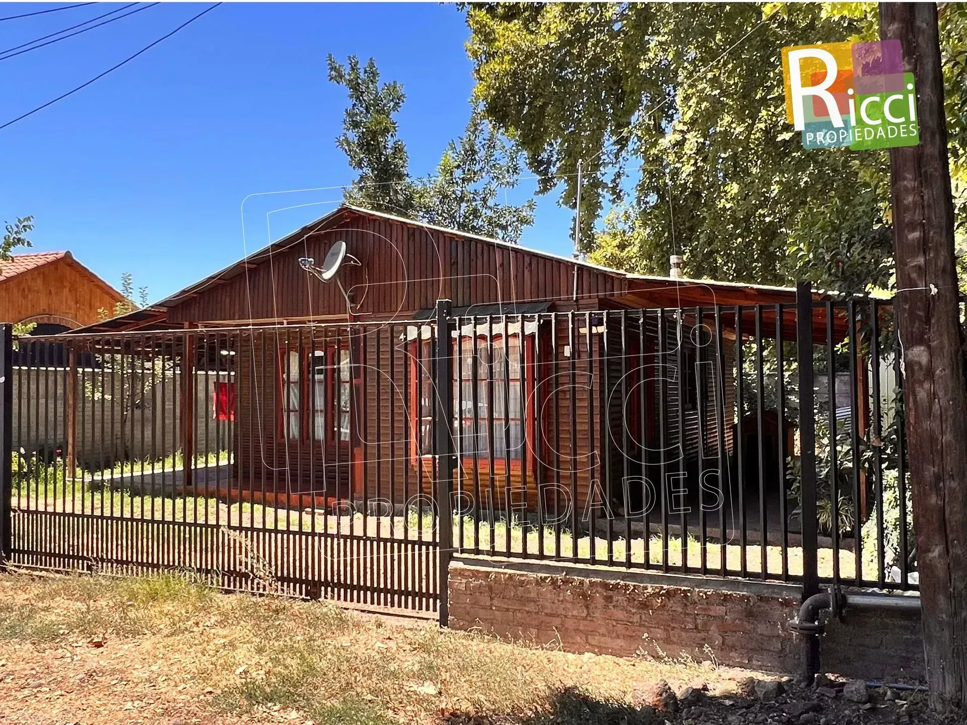 Casa rústica de madera detrás de una valla metálica negra, bajo los árboles y un cielo azul.