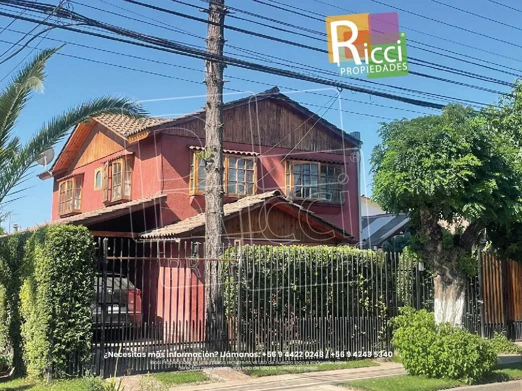 Casa roja de dos plantas con una valla metálica negra y abundante vegetación bajo un cielo azul