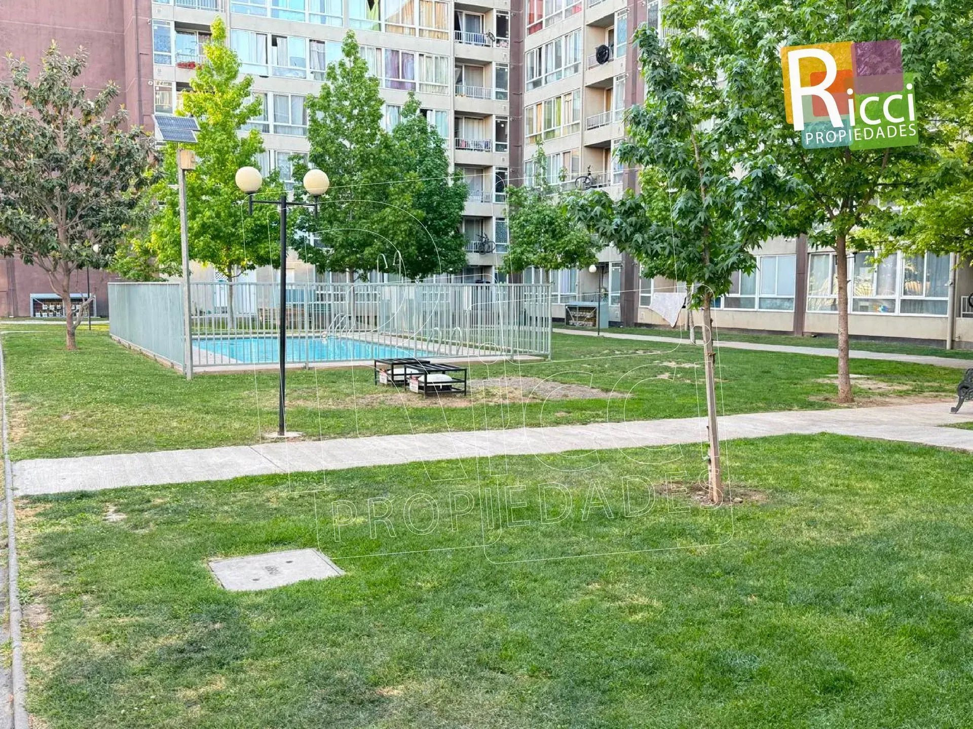 Patio comunitario con césped, árboles, bancos y un parque infantil vallado junto a edificios residenciales