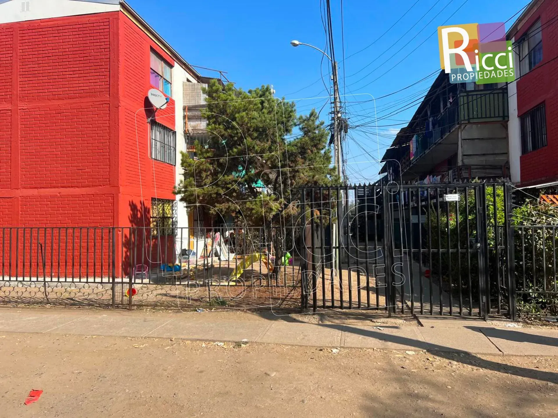 Vista desde la calle de un edificio rojo junto a una puerta negra bajo un cielo azul, con un letrero de “Ricci” al fondo