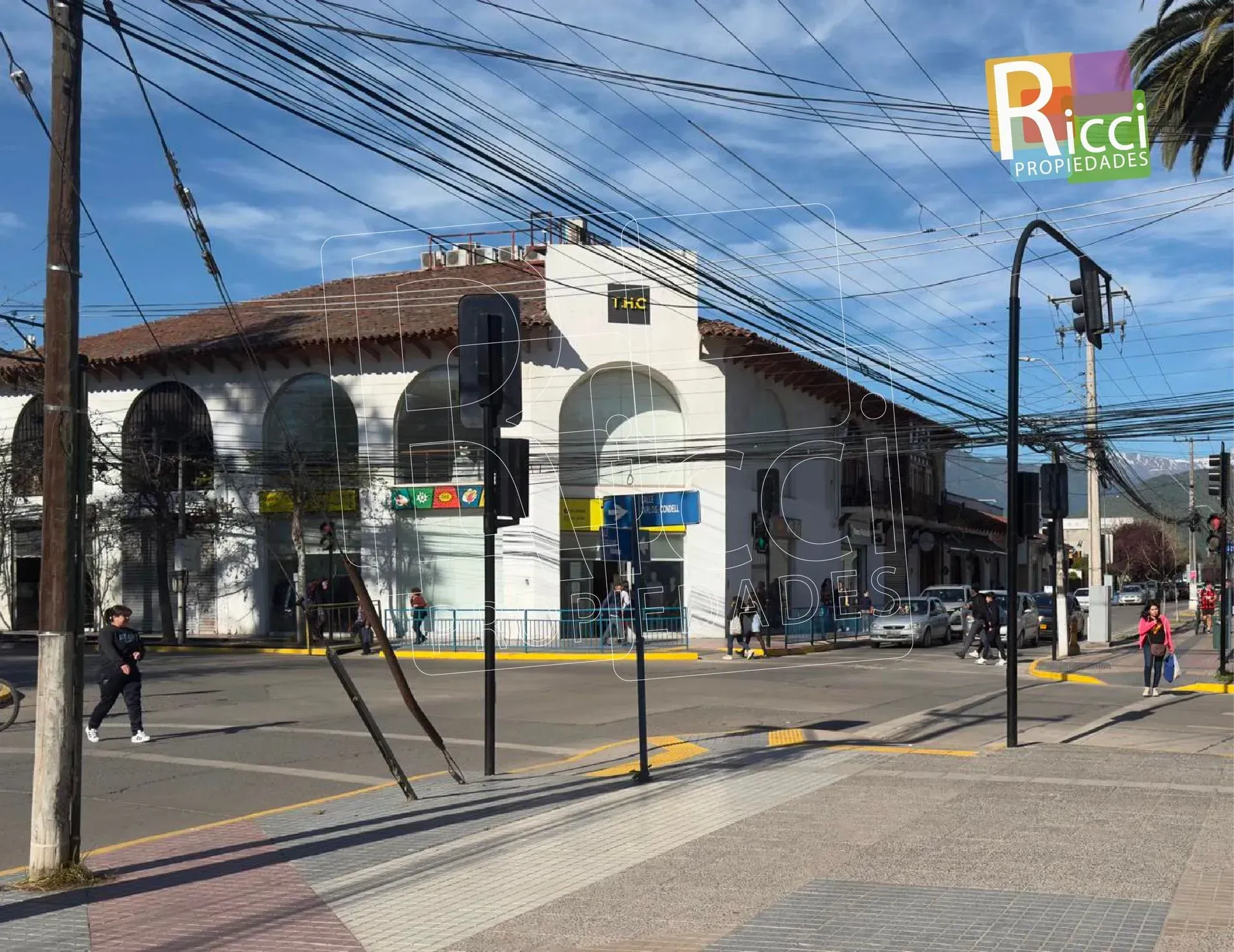 Esquina con un edificio blanco con arcos, vías de tranvía, cables aéreos y algunos peatones bajo un cielo azul