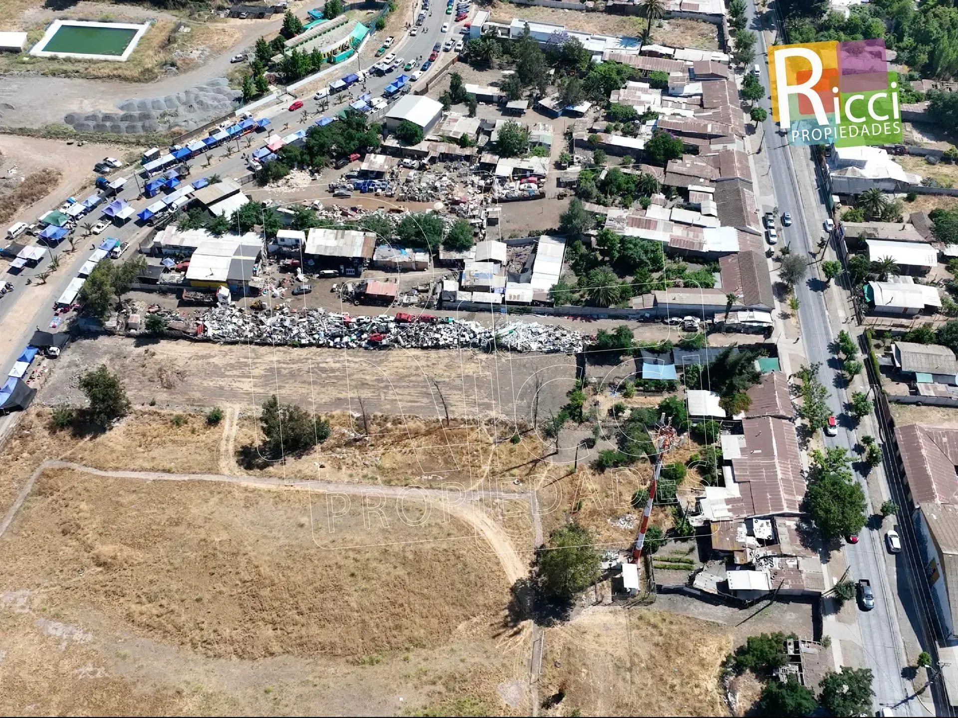 Calle estrecha con una valla de chapa ondulada, casas desgastadas y un coche rojo bajo un cielo azul despejado