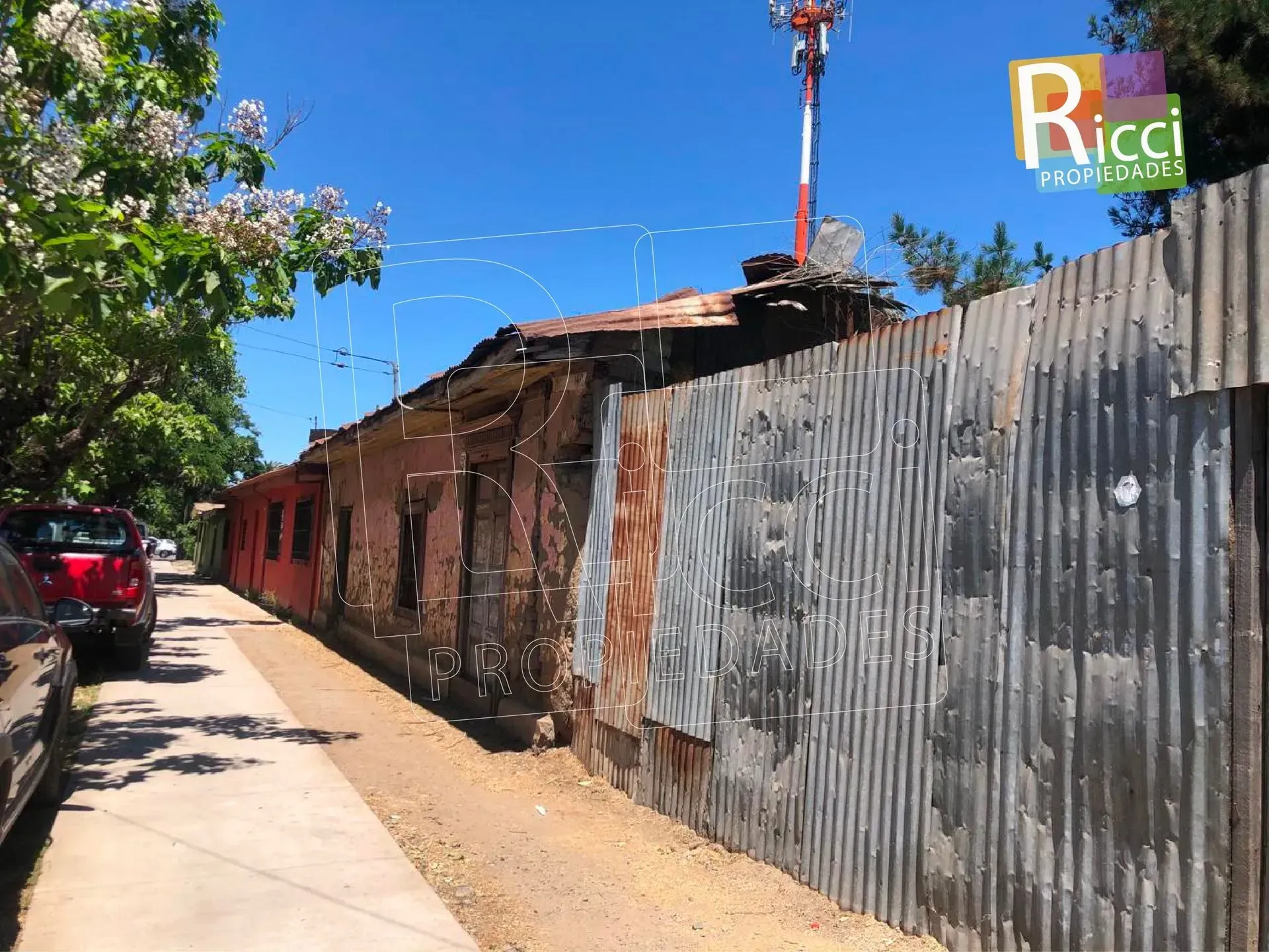 Calle estrecha con una valla de chapa ondulada, casas desgastadas y un coche rojo bajo un cielo azul despejado