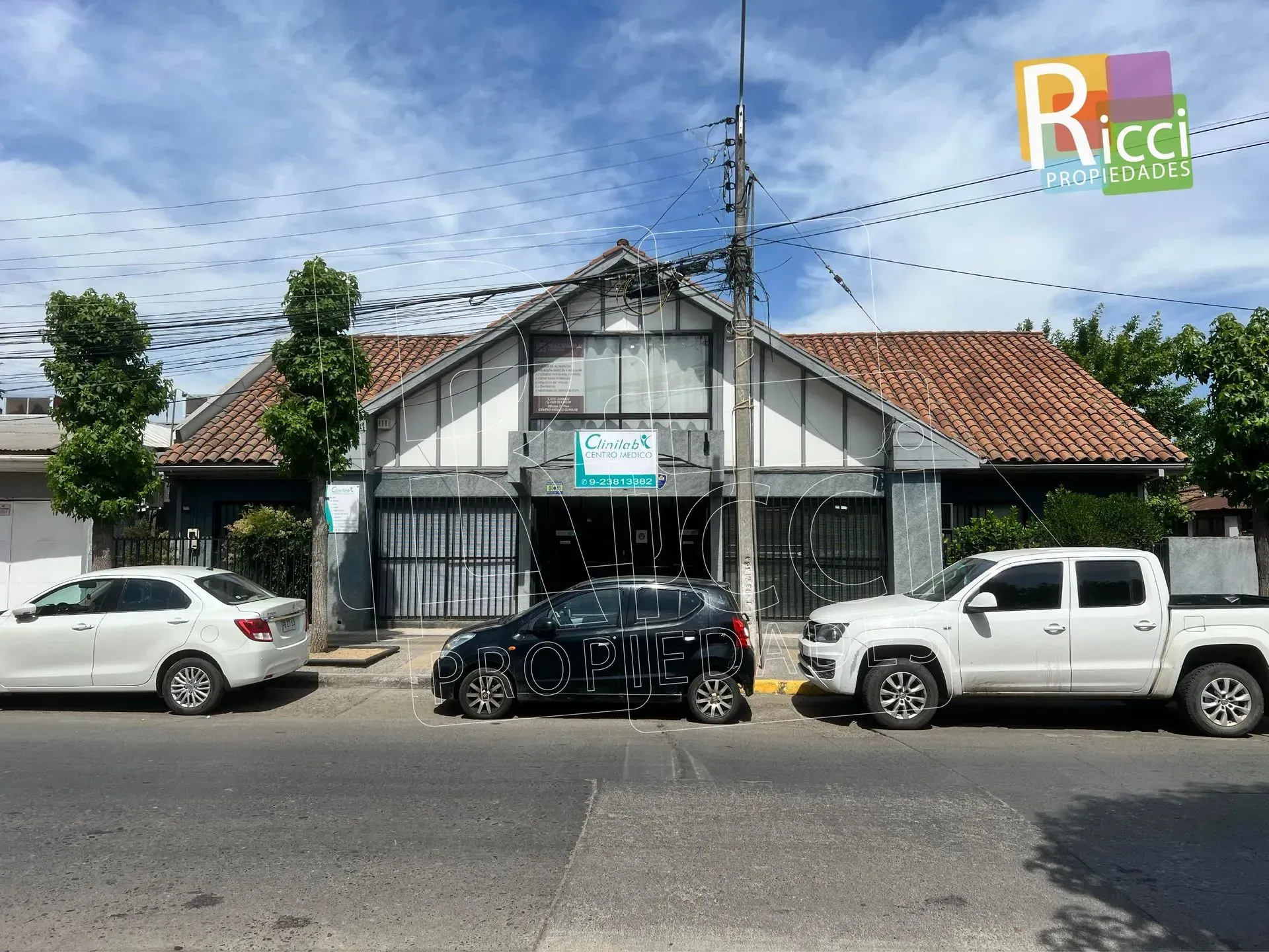 Vista de una pequeña tienda a pie de calle, con coches aparcados y un tejado de tejas bajo un cielo nublado