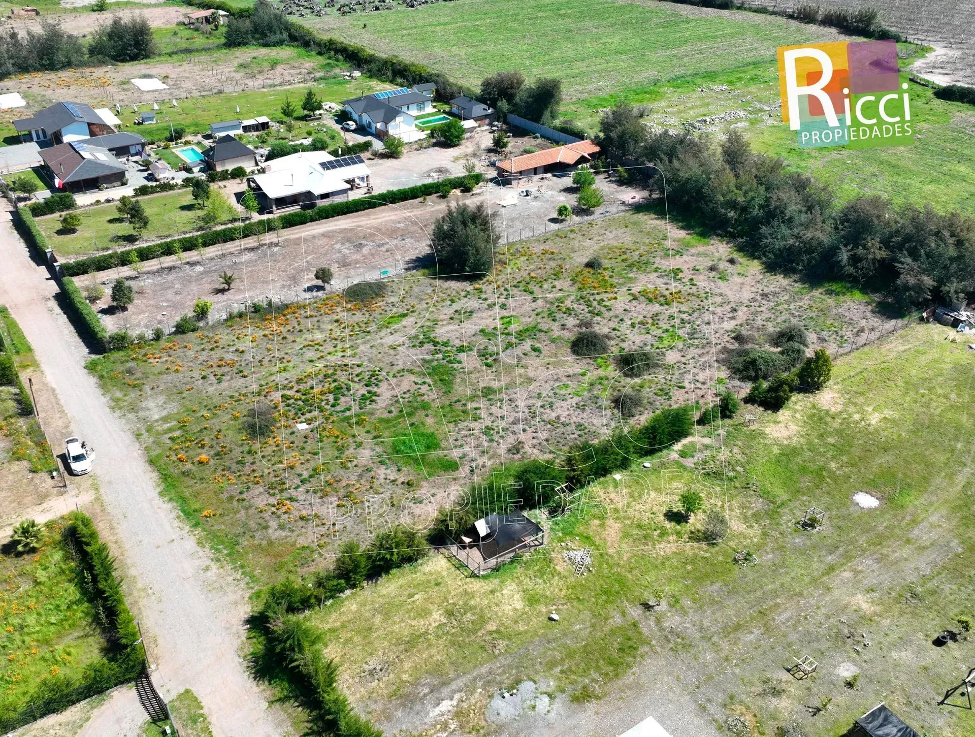 Vista aérea de un terreno baldío cubierto de maleza junto a una carretera, con casas y árboles cerca.