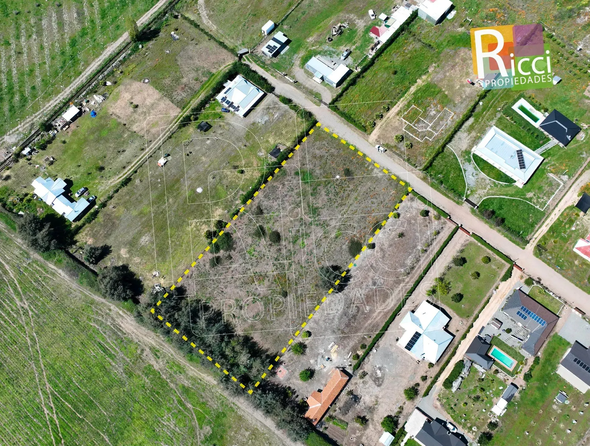 Vista aérea de un terreno baldío cubierto de maleza junto a una carretera, con casas y árboles cerca.