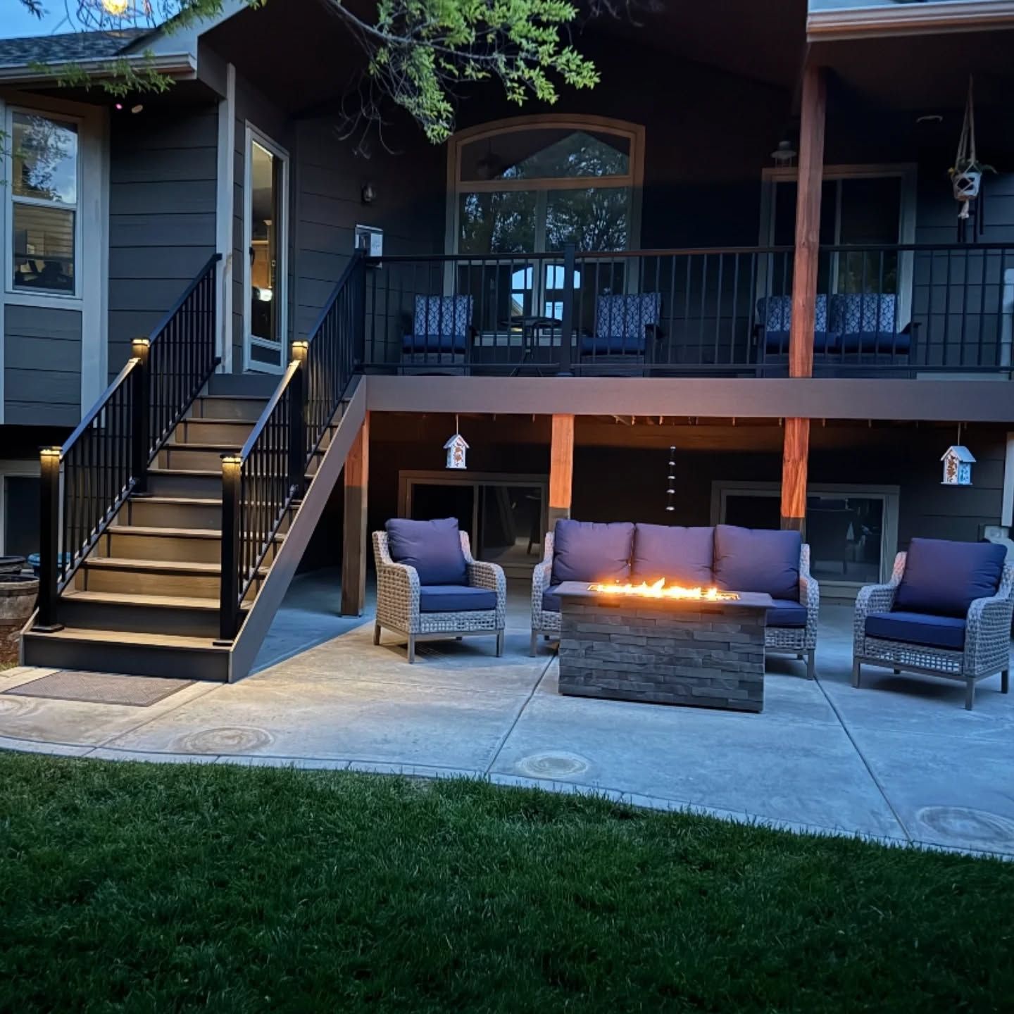 A backyard patio at dusk with a lit fire pit, a sofa, two chairs, and a staircase leading to a wooden deck above.