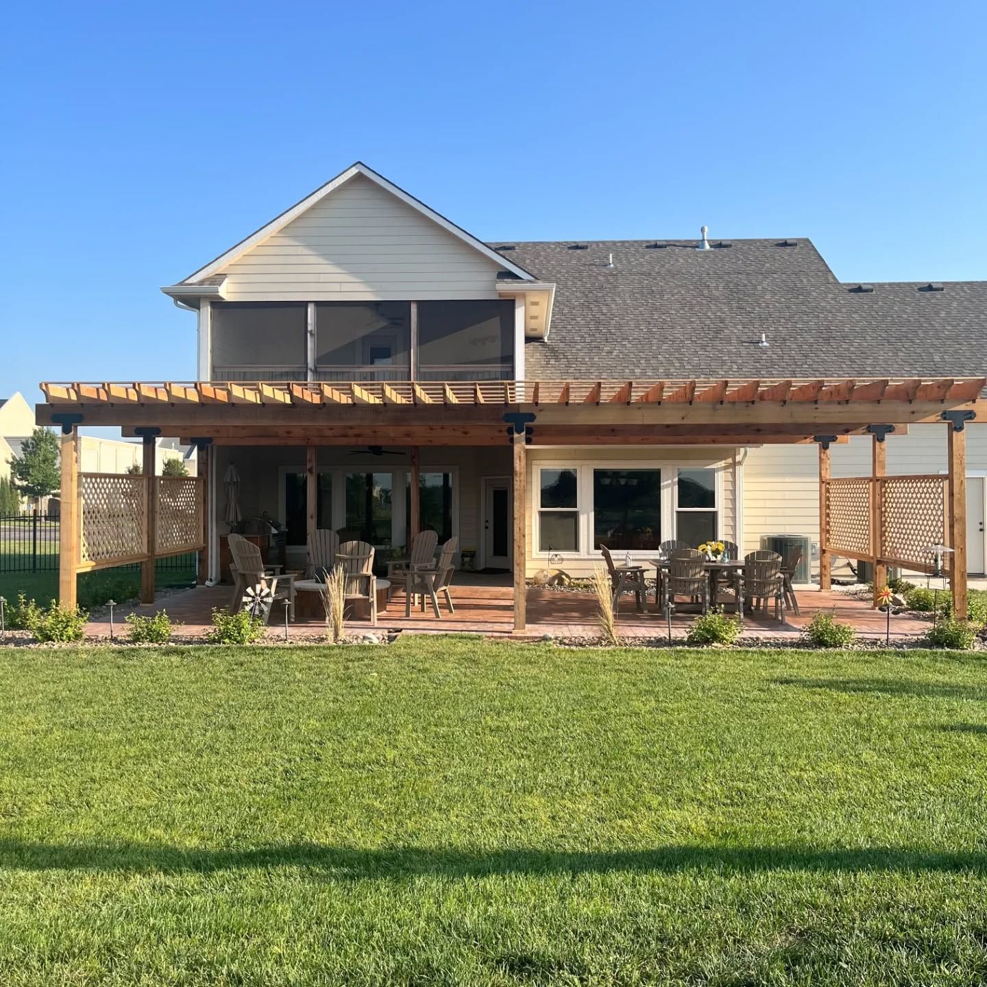 A two-story house with a large wooden pergola covering a patio area, set against a green lawn under a clear blue sky.