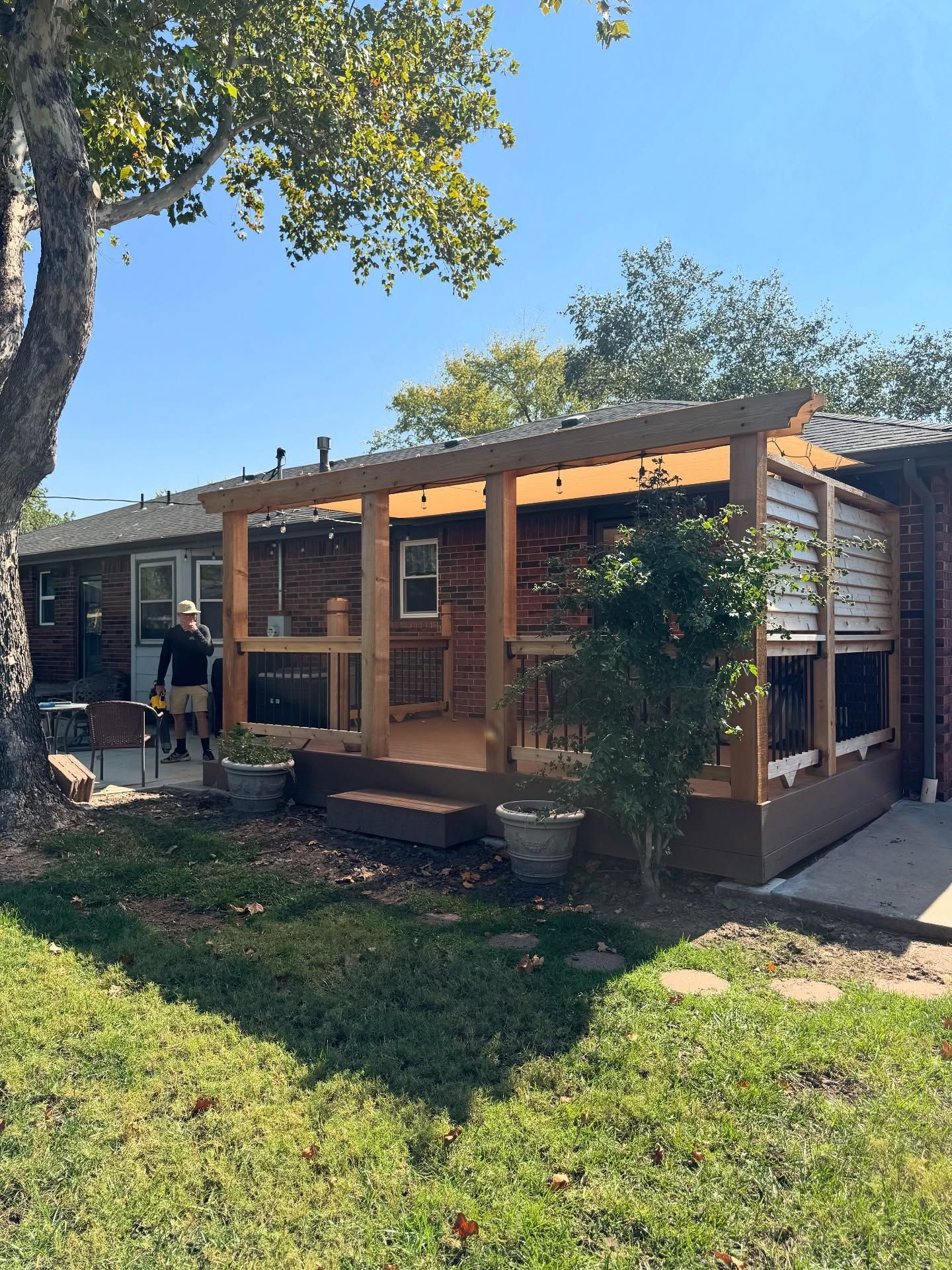 A wooden pergola covers an outdoor deck attached to the back of a brick house on a sunny day.
