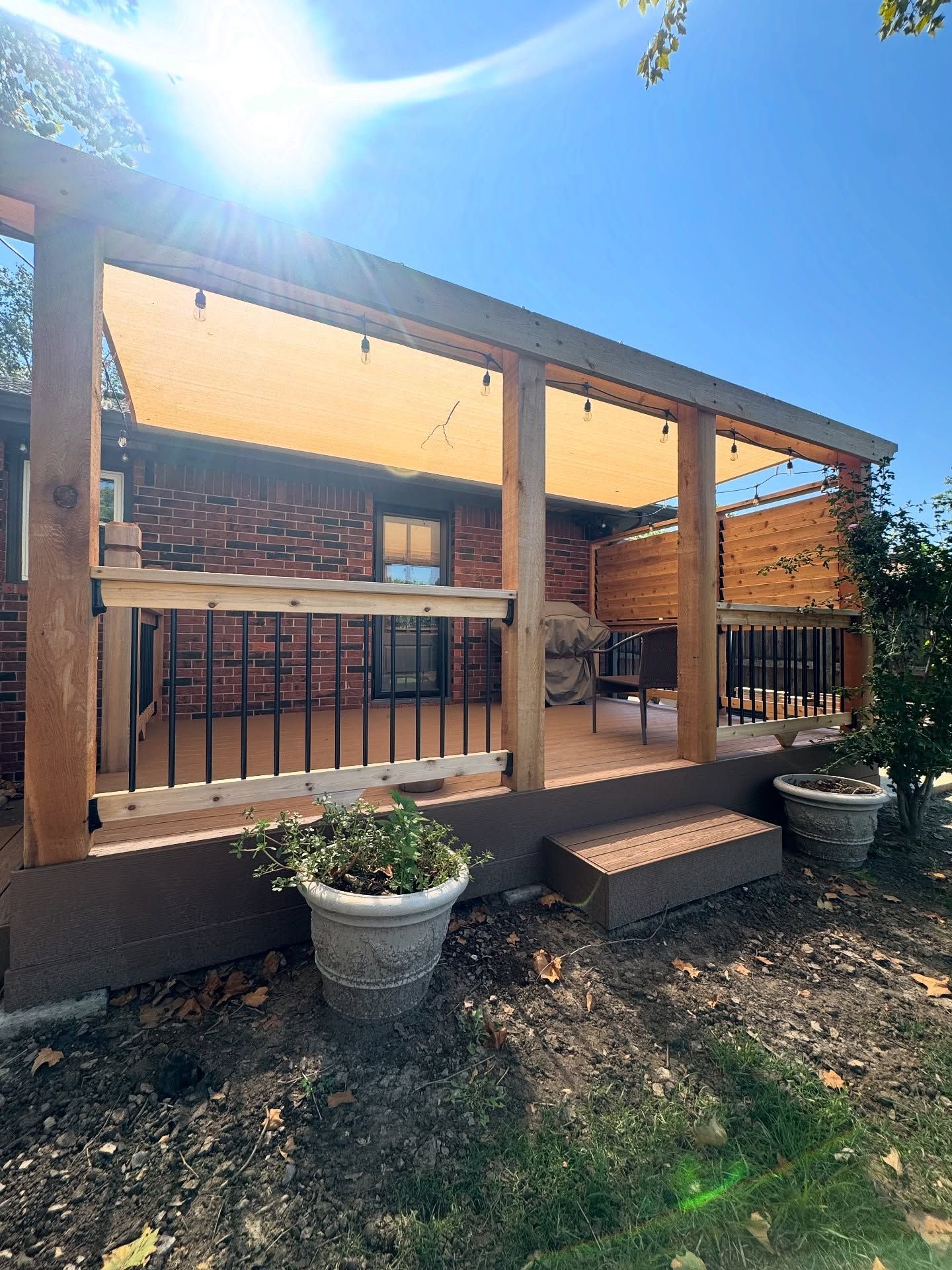 A wooden deck with a tan sun shade, black railings, and potted plants attached to a brick house under a blue sky.