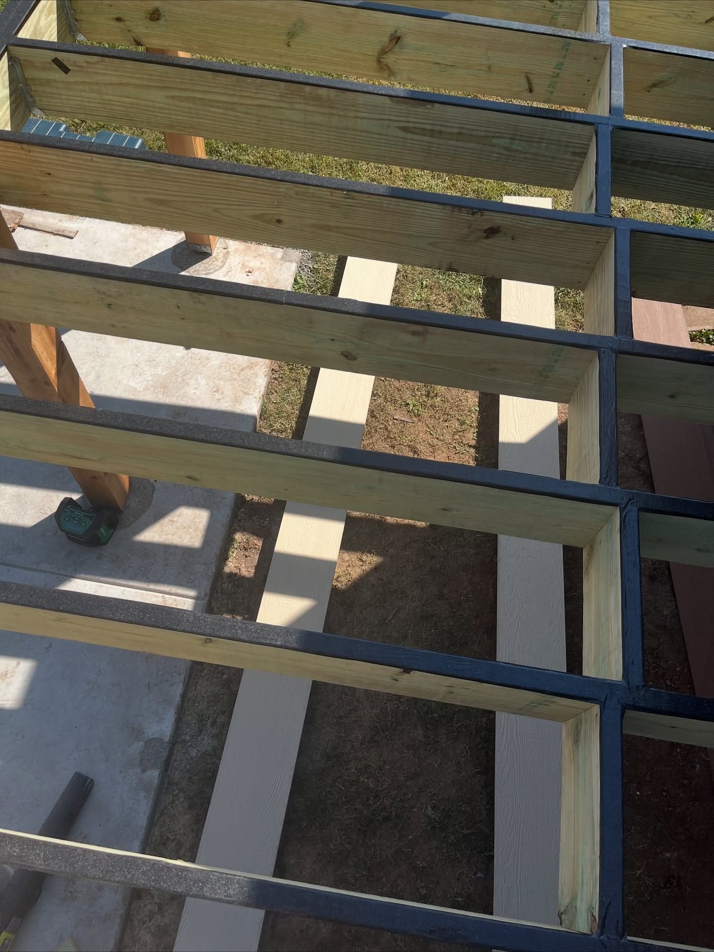 Wooden deck joists in a grid pattern, partially painted with black protective tape along the top edges.