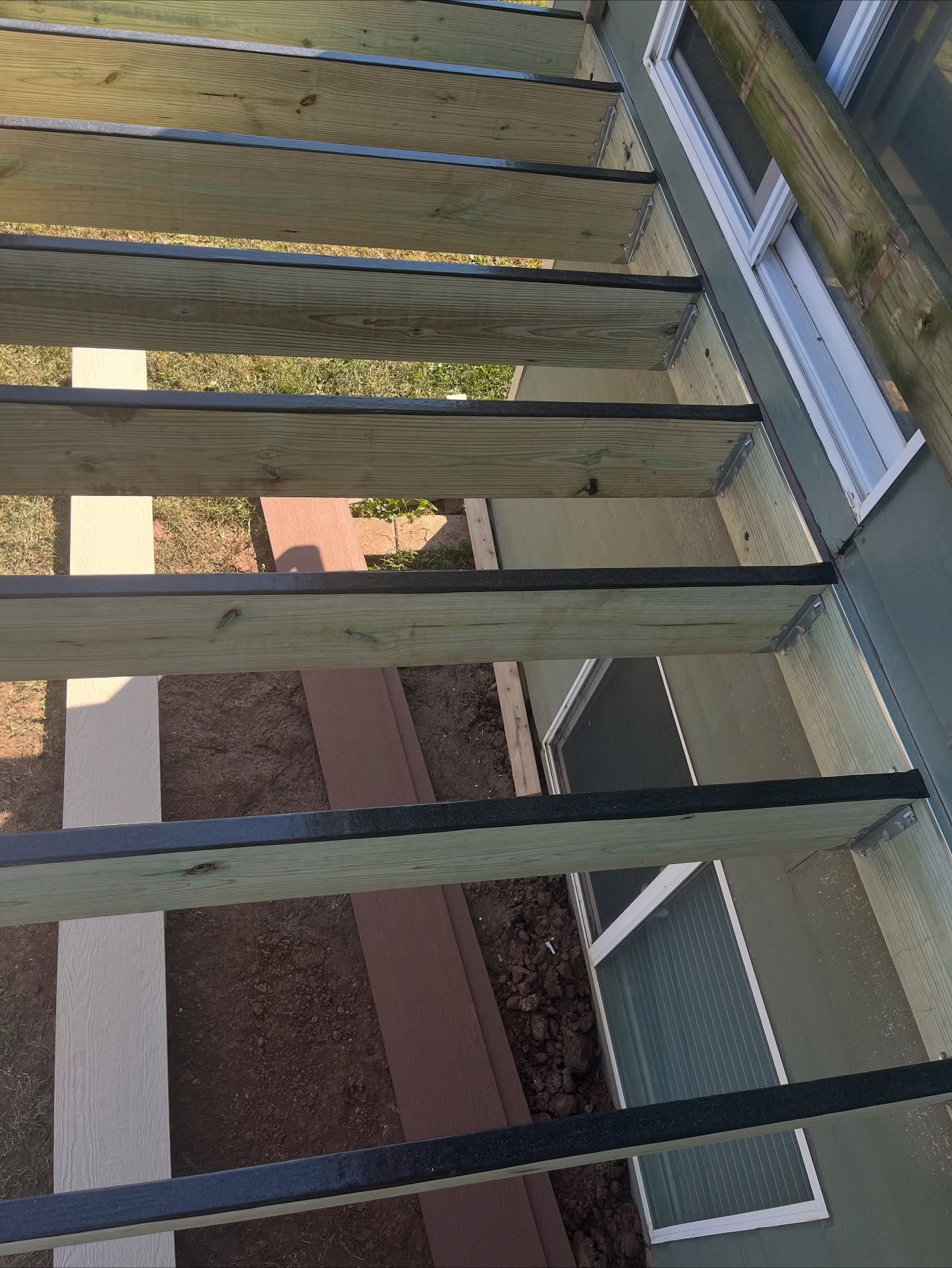 High-angle view of a wooden deck frame under construction, with joists attached to a house wall above a window well.