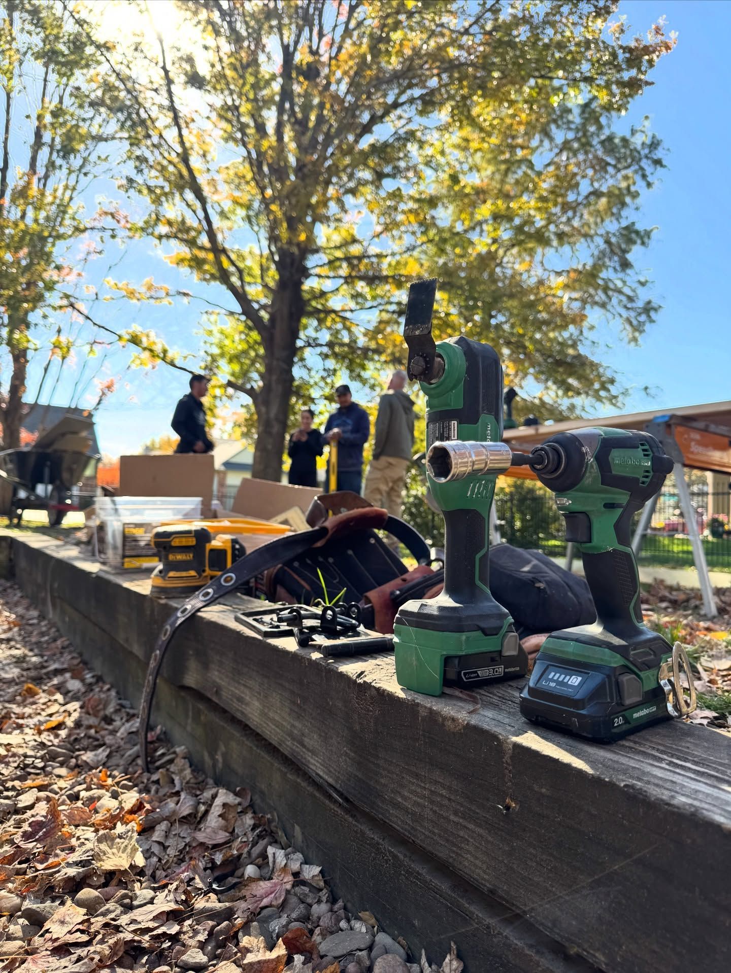 Two power drills sit on a wooden beam outdoors, with blurred figures working in the background on a sunny day.