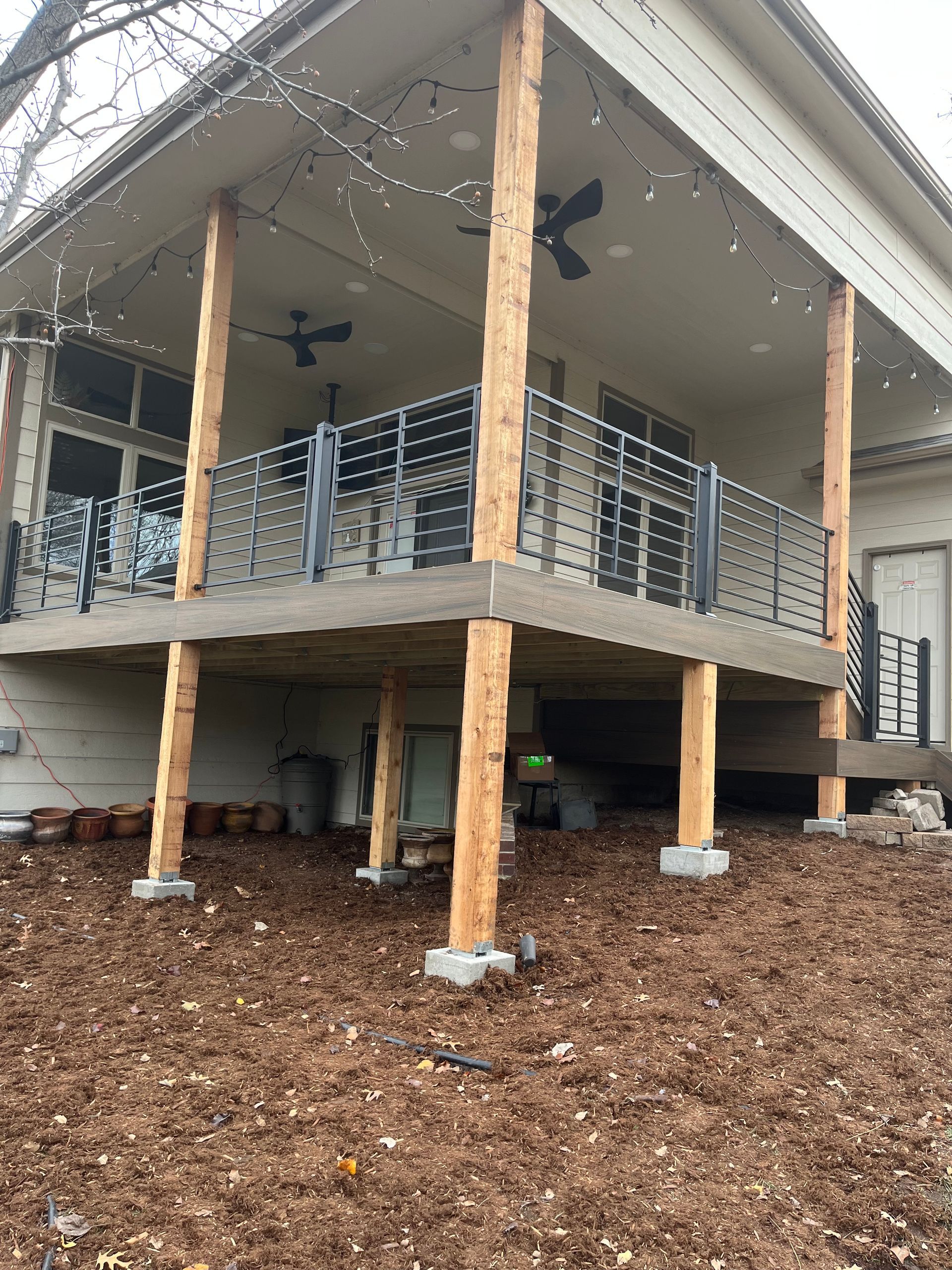 An elevated outdoor patio with a metal railing, supported by rustic wooden posts resting on concrete footings.
