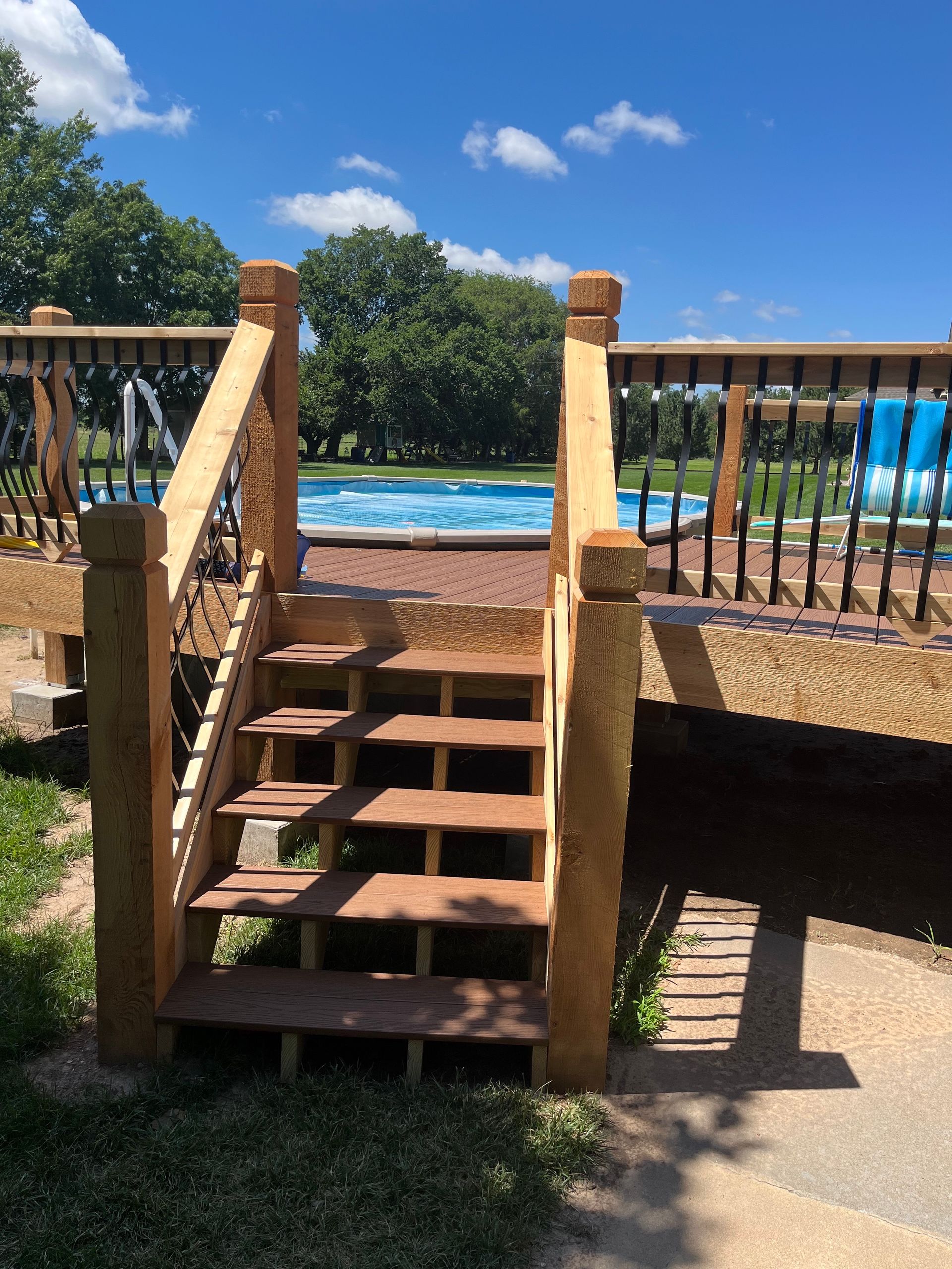 Wooden steps leading up to a deck with railings, overlooking a backyard swimming pool under a sunny blue sky.