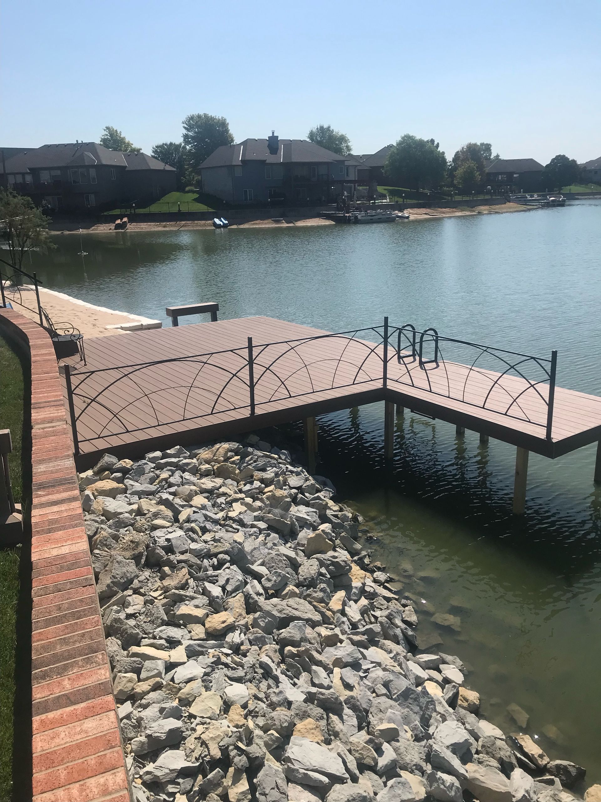 A brown composite deck with black metal railings extends over a lake, fronted by a rocky shoreline and a brick retaining wall.