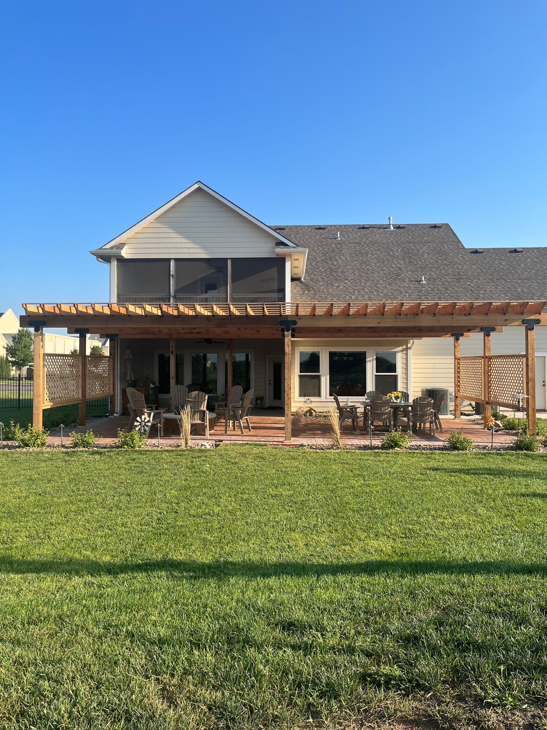 A beige house with a large wooden pergola covering a stone patio, viewed from a grassy backyard under a clear blue sky.