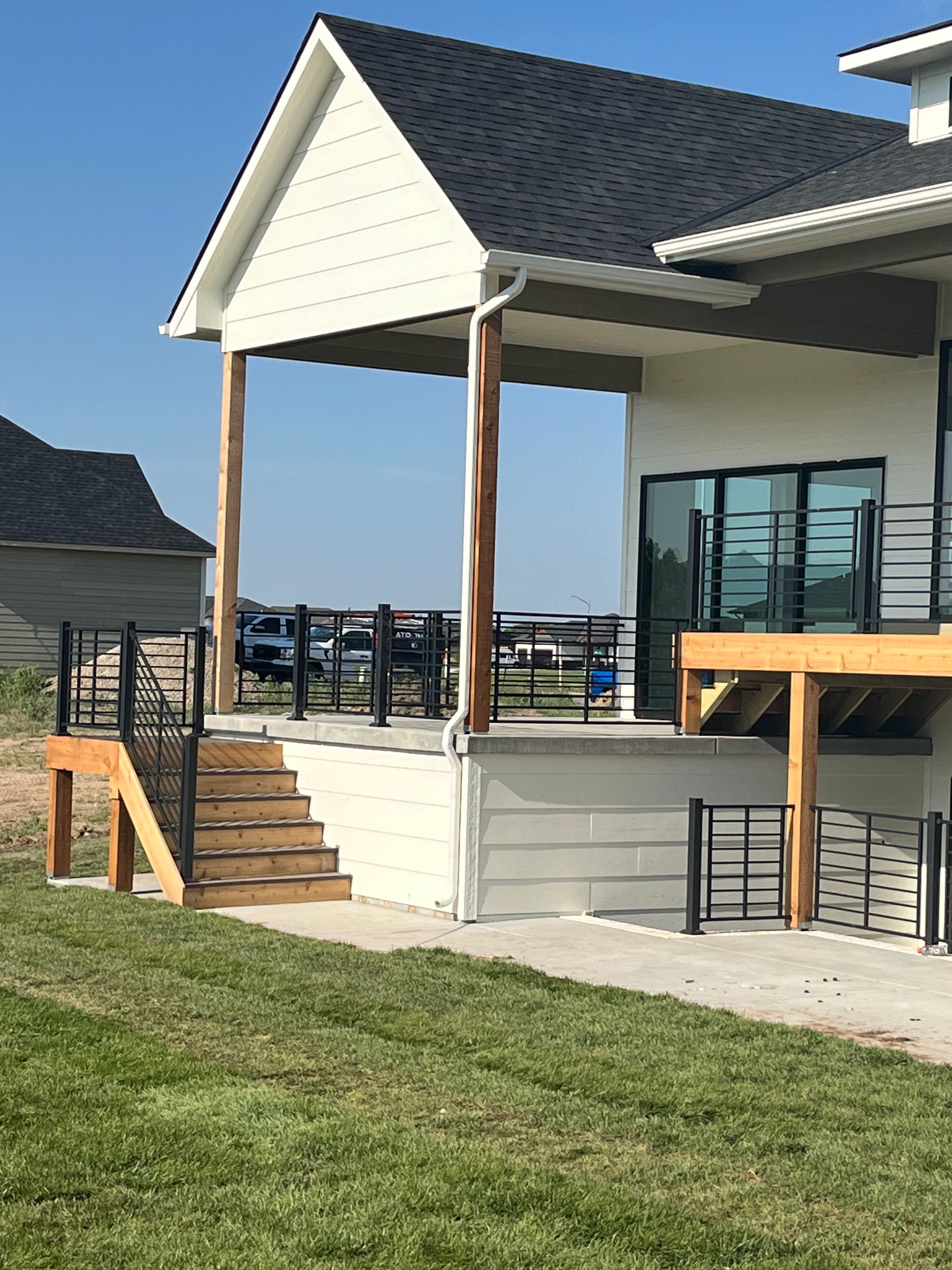 A covered back patio with a wooden staircase and metal railings attached to a white modern home with a black roof.