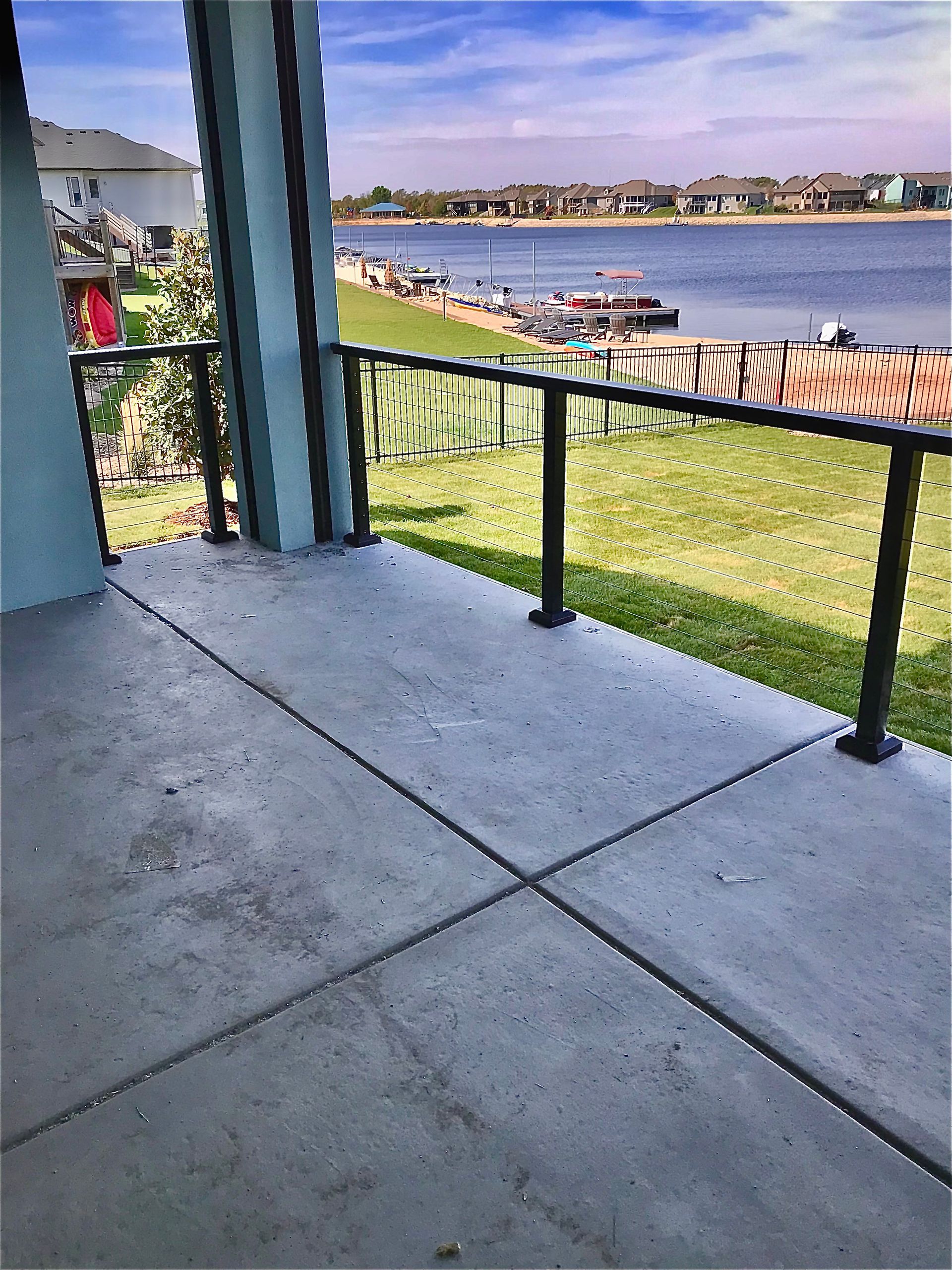Concrete patio with black metal railing overlooking a lakefront neighborhood on a sunny day.