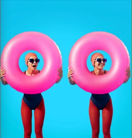 Two women in swimsuits are wearing woow sunglasses and
holding pink inner tubes over their heads.