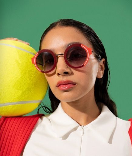 A woman wearing woow sunglasses is holding a tennis ball on her shoulder.