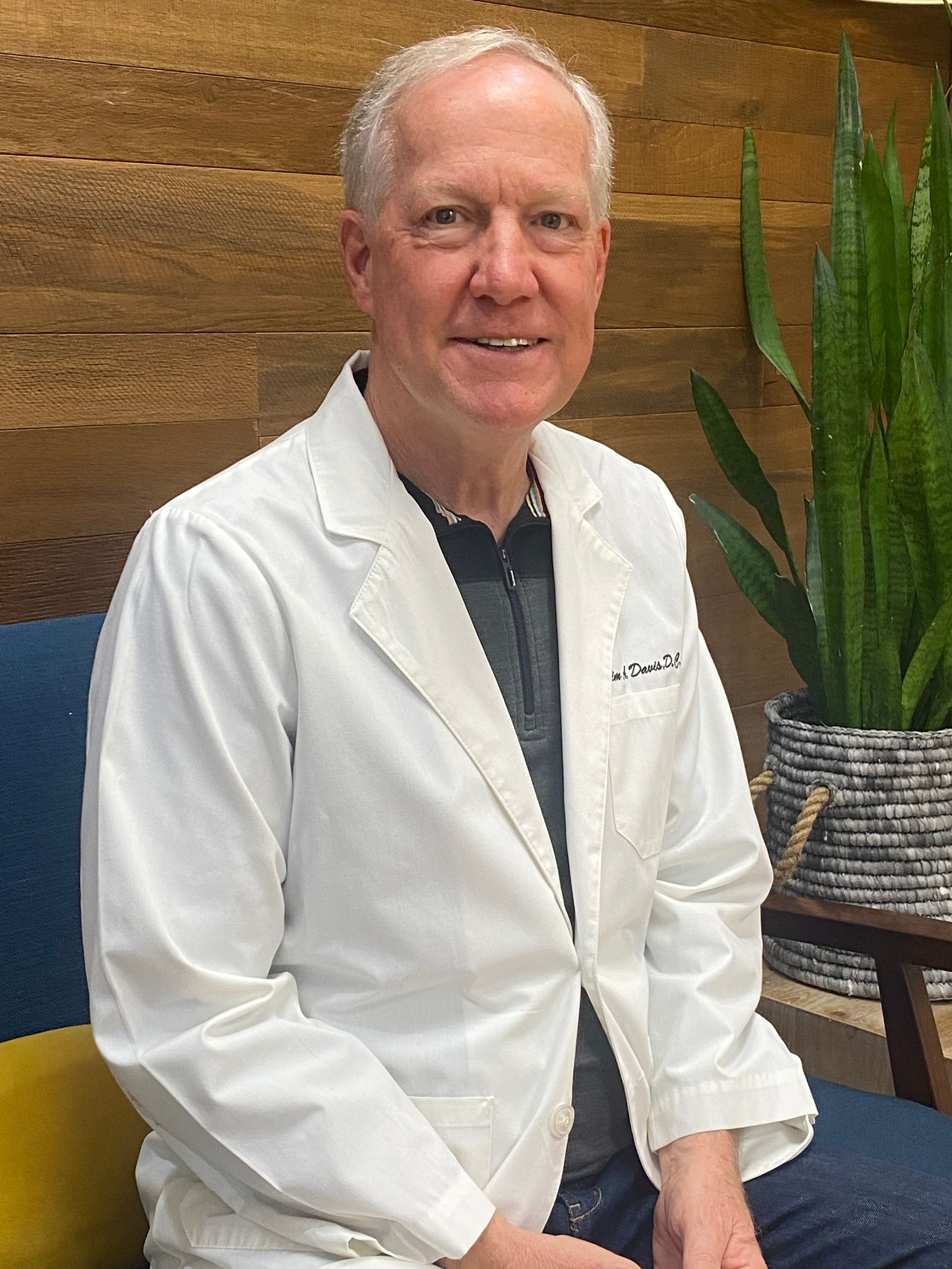 Man in white lab coat smiles, seated near a plant and wood-paneled wall.