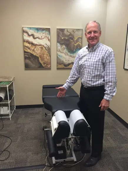 Man in button-down shirt points to a chiropractic table in a doctor's office. Artwork on the wall.