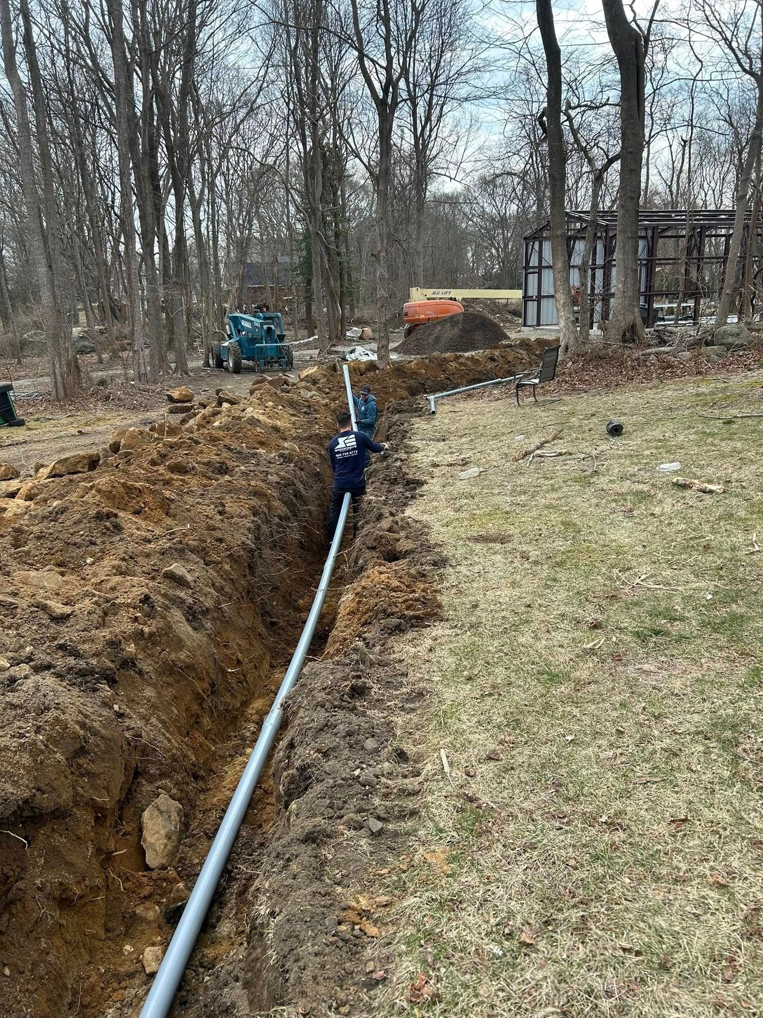 A trench with a pipe running through it in a wooded area, with construction equipment visible.