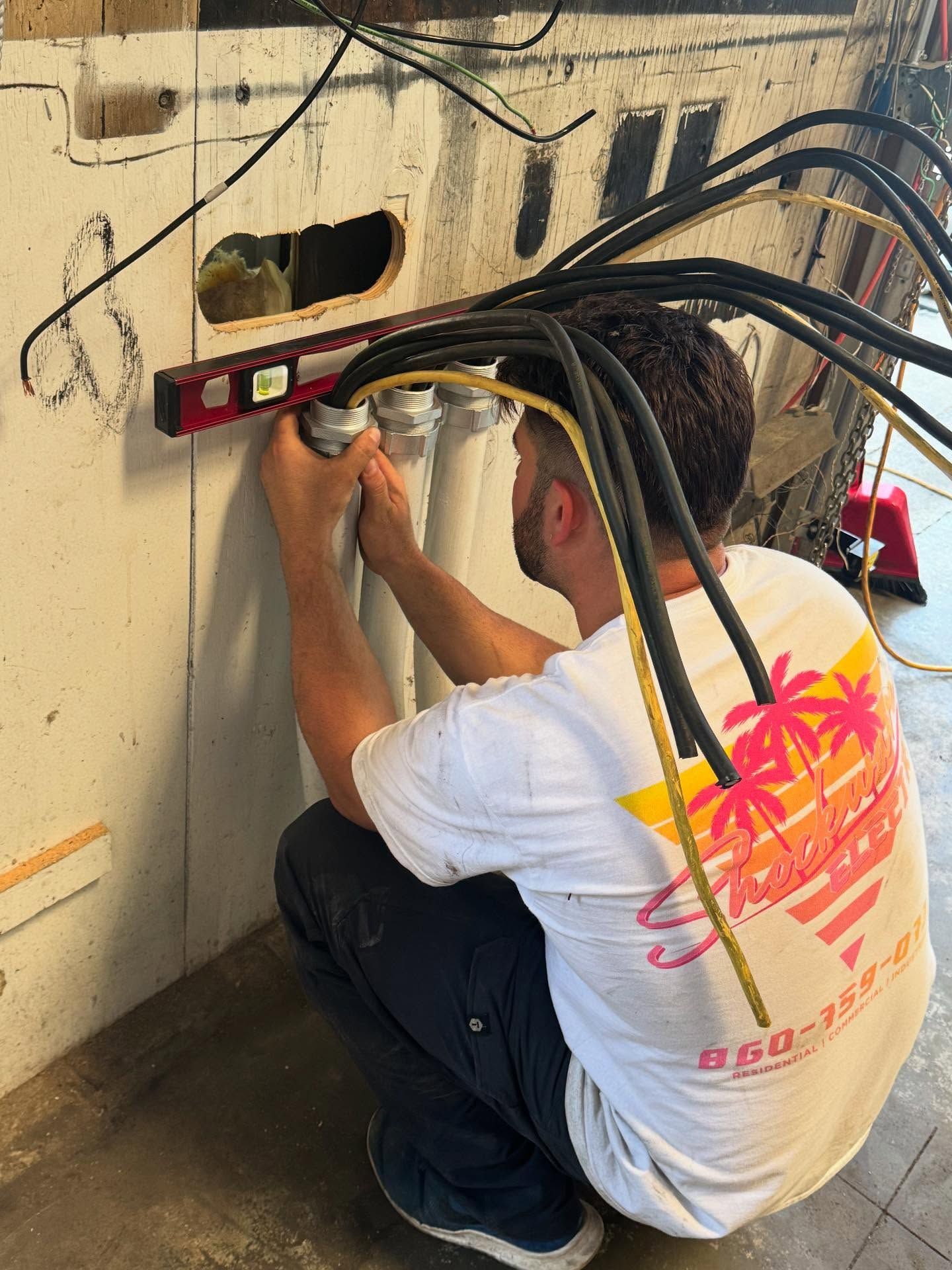Man using a level on electrical conduit. He's wearing a white shirt with a tropical design and kneeling. 