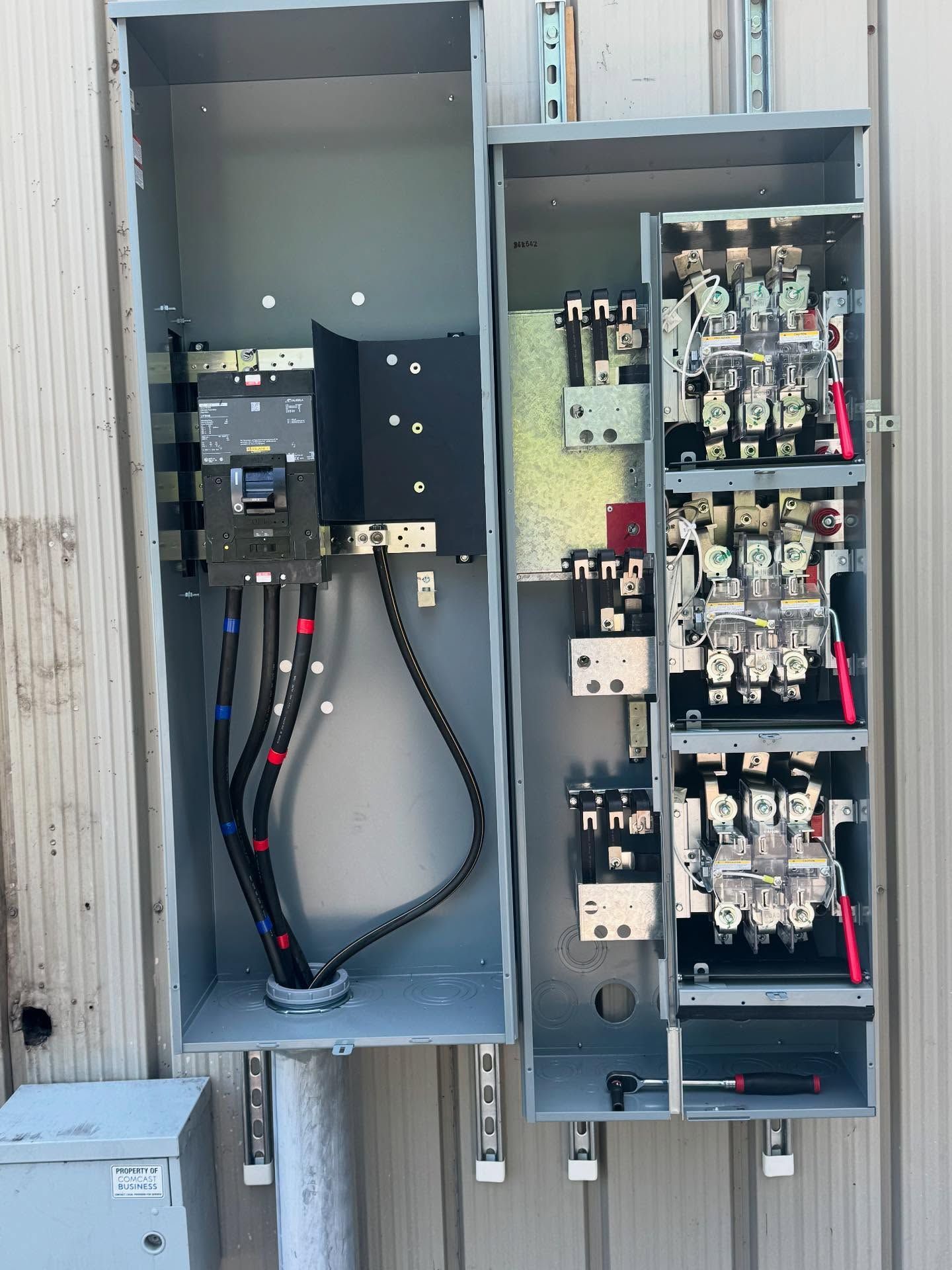 Metal electrical panel with open compartments, wires, and circuit breakers. Mounted on a corrugated metal wall.