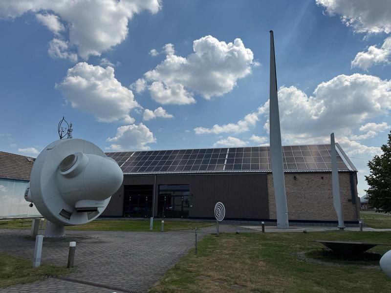 Modernes Gebäude mit Solarpaneelen unter blauem Himmel, neben einer weißen abstrakten Skulptur.